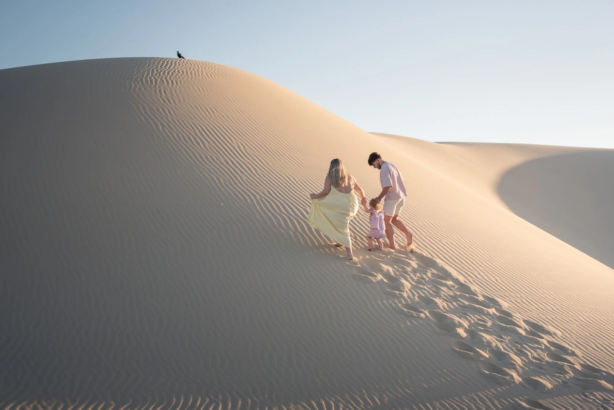 Family of three walking hand-in-hand on sand dunes in a desert with clear sky.