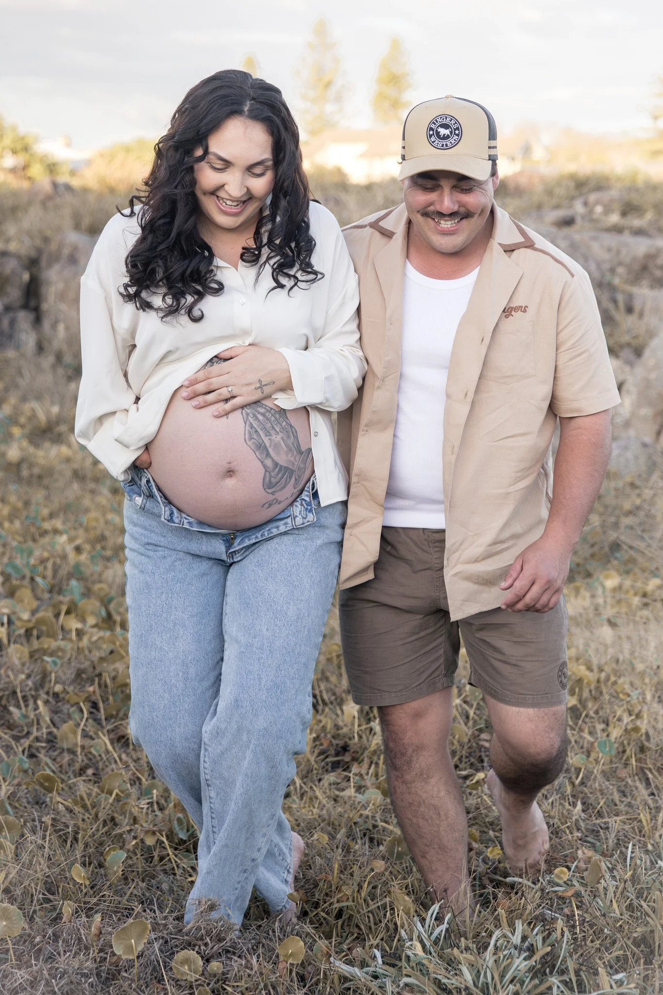 A couple walking outside in a field of dry grass and plants, smiling, with the woman showing her pregnant belly and tattoos on her hand.