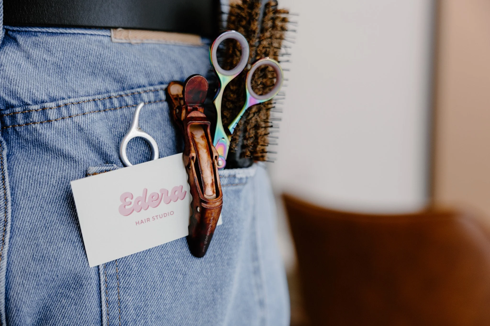 Close-up of a person's waist wearing blue denim jeans with hair salon tools and a business card tucked in the pocket, including rainbow-colored scissors, a brown hair clip, and a wooden comb.