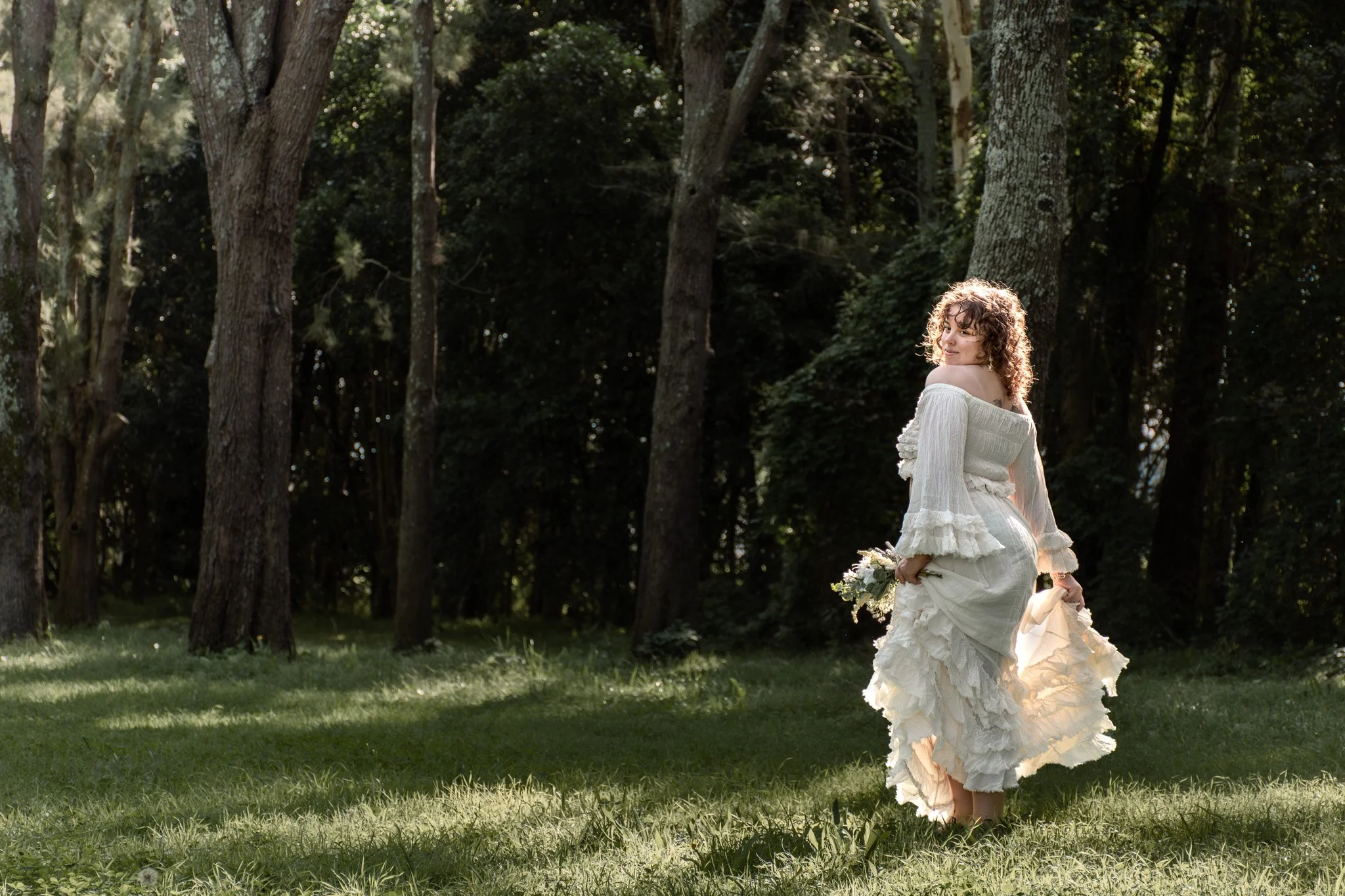 A woman in a white dress holding a bouquet of flowers standing outdoors in a wooded area with sunlight filtering through trees.