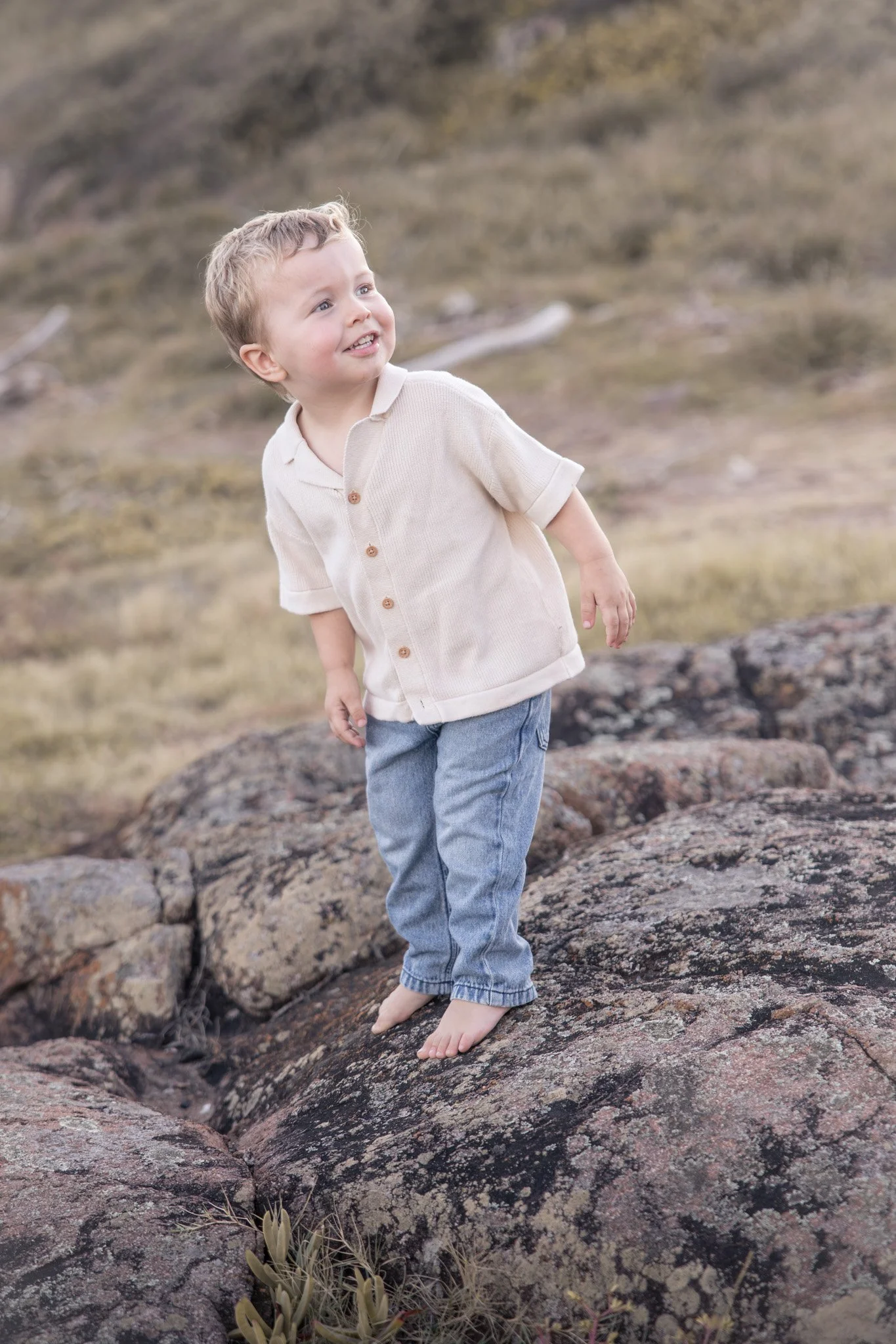 A young boy with blonde hair and a beige polo shirt standing barefoot on rocks in an outdoor natural environment with a rocky background and sparse vegetation.