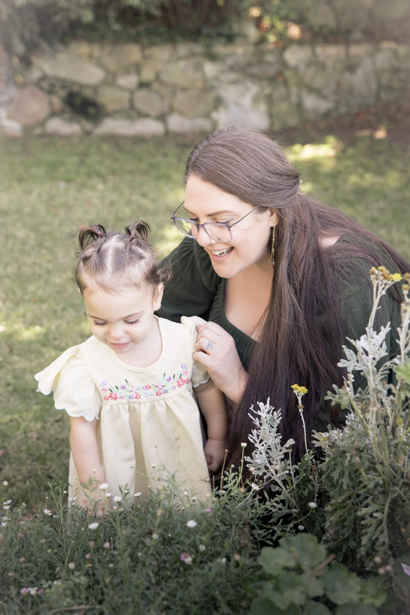 A woman with long brown hair, glasses, and earrings laughs while crouching Next to a young girl with light brown hair styled in pigtails, wearing a yellow dress with floral embroidery, in a garden with grass and small flowers.