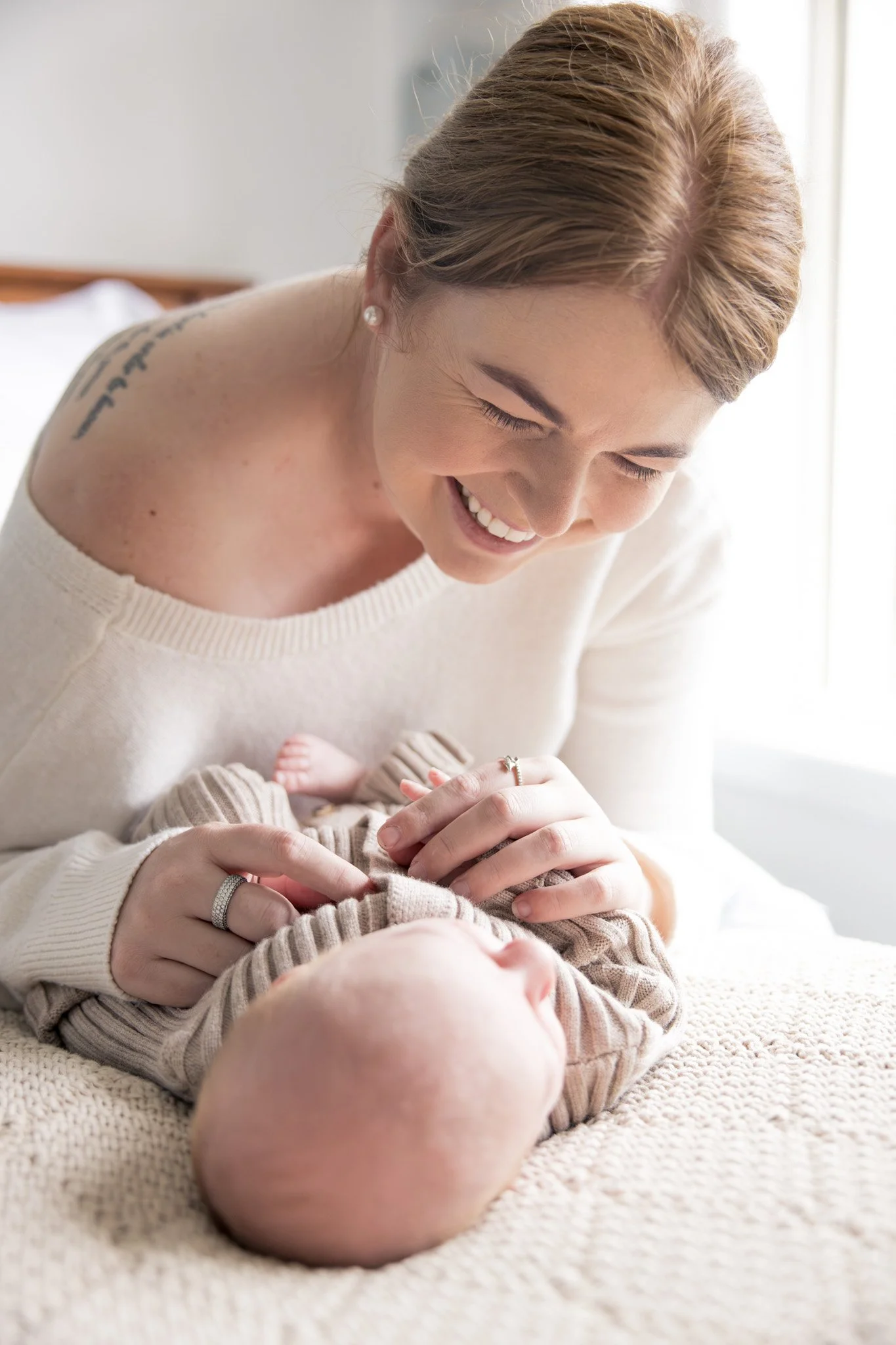 A woman with light brown hair and pearl earrings smiling down at a baby lying on a cozy beige blanket. The woman is wearing a white sweater, and the baby is dressed in beige and brown striped clothing. The scene appears to be in a bright room with na