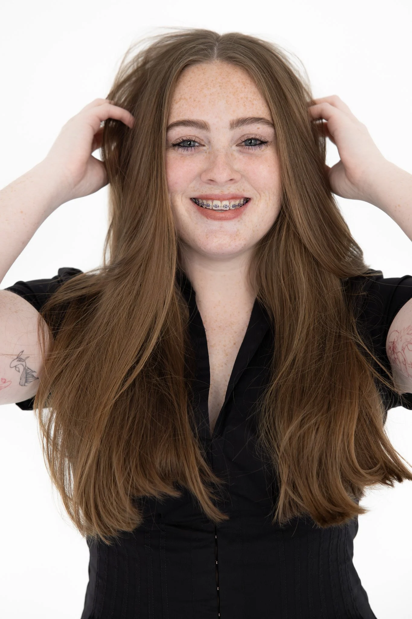 A young woman with long red hair, freckles, and blue eyes, smiling with braces. She is wearing a black top and is touching her hair with both hands against a plain white background. Taken at sister studio newcastle