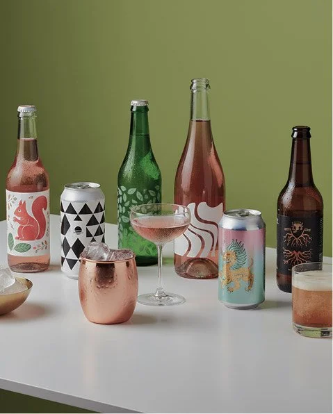Various bottles, cans, and glasses of beverages on a white table against a green background, including beer bottles, a cocktail glass, and metallic cups.