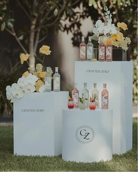 Display of colorful crafted beverages in bottles with floral decorations on three white podiums outdoors