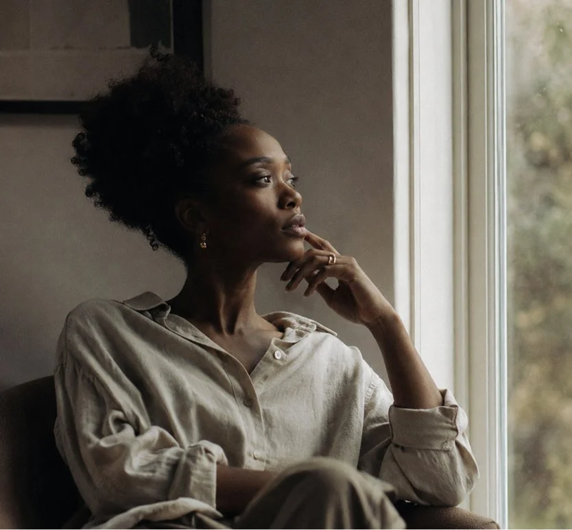A woman with dark curly hair sitting by a window, gazing outside thoughtfully.