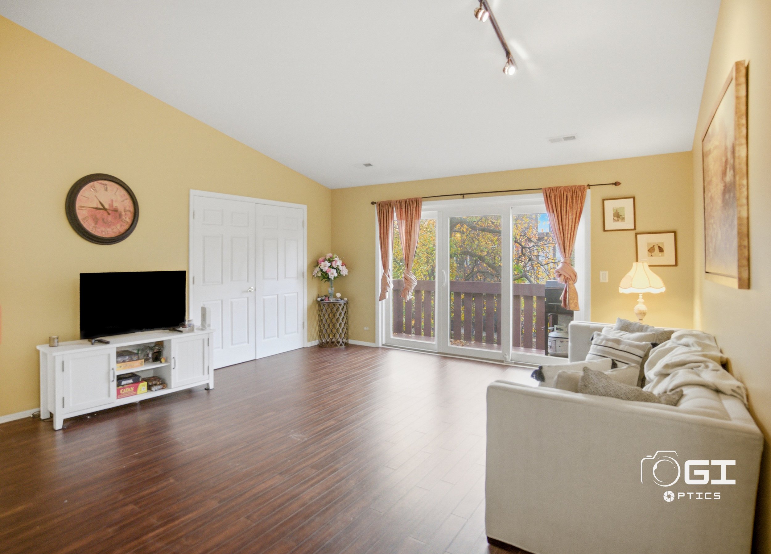 Living room with yellow walls, white sofa with pillows, wooden floor, sliding glass door with pink curtains leading to balcony with trees outside, small white TV stand with TV, wall clock, and framed artwork.