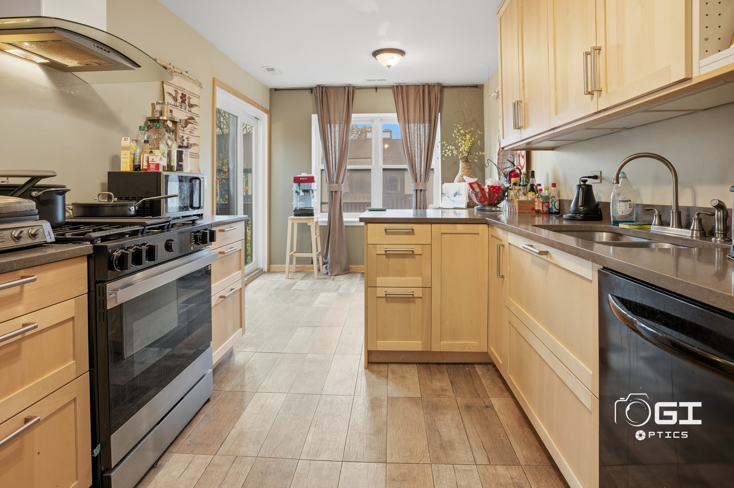 Kitchen with wooden cabinets, stainless steel appliances, and a window with brown curtains, cluttered with bottles and kitchen utensils.