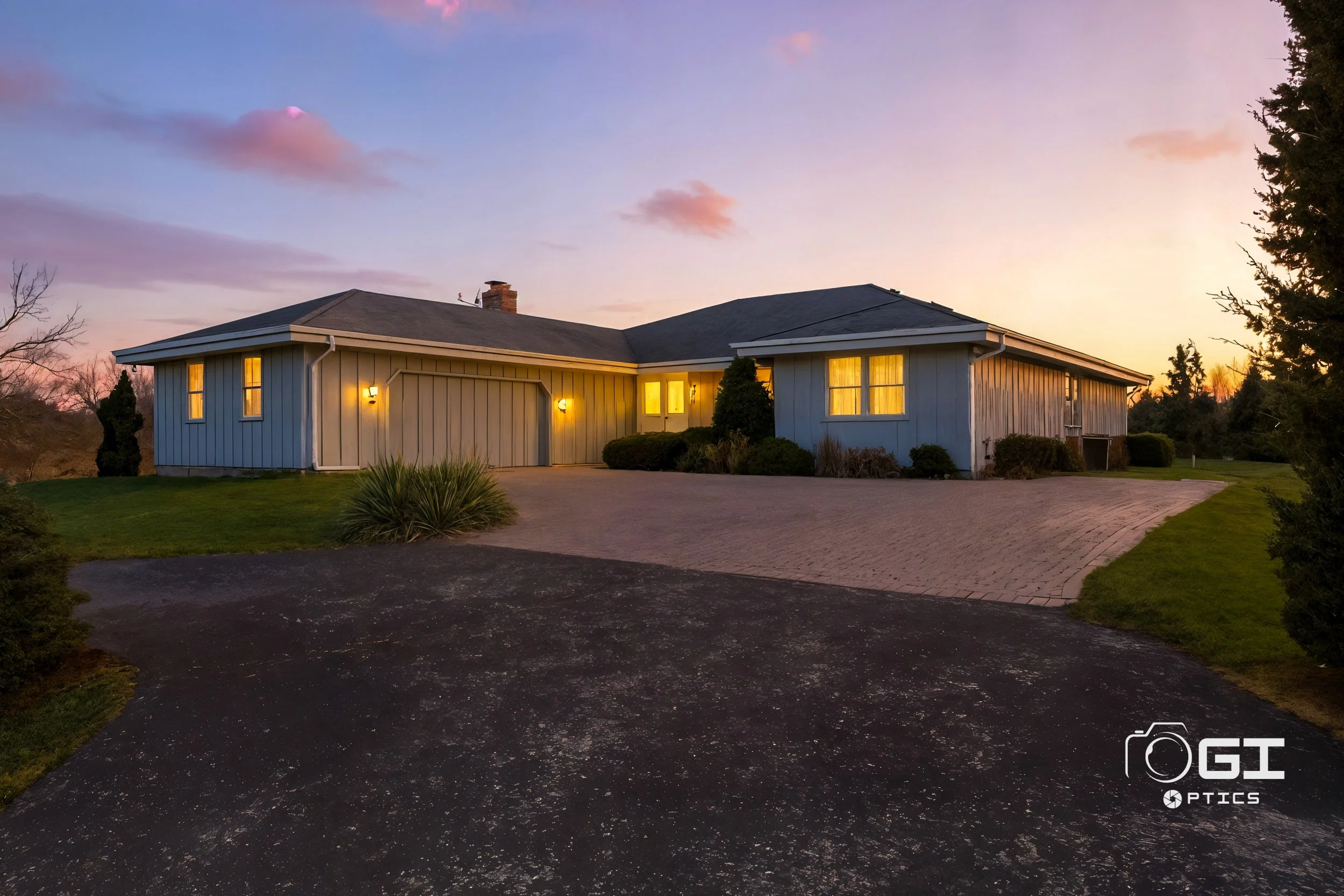 A single-story house at sunset with illuminated windows and exterior lights, surrounded by a landscaped yard with bushes and trees, a paved driveway, and a partly cloudy sky.