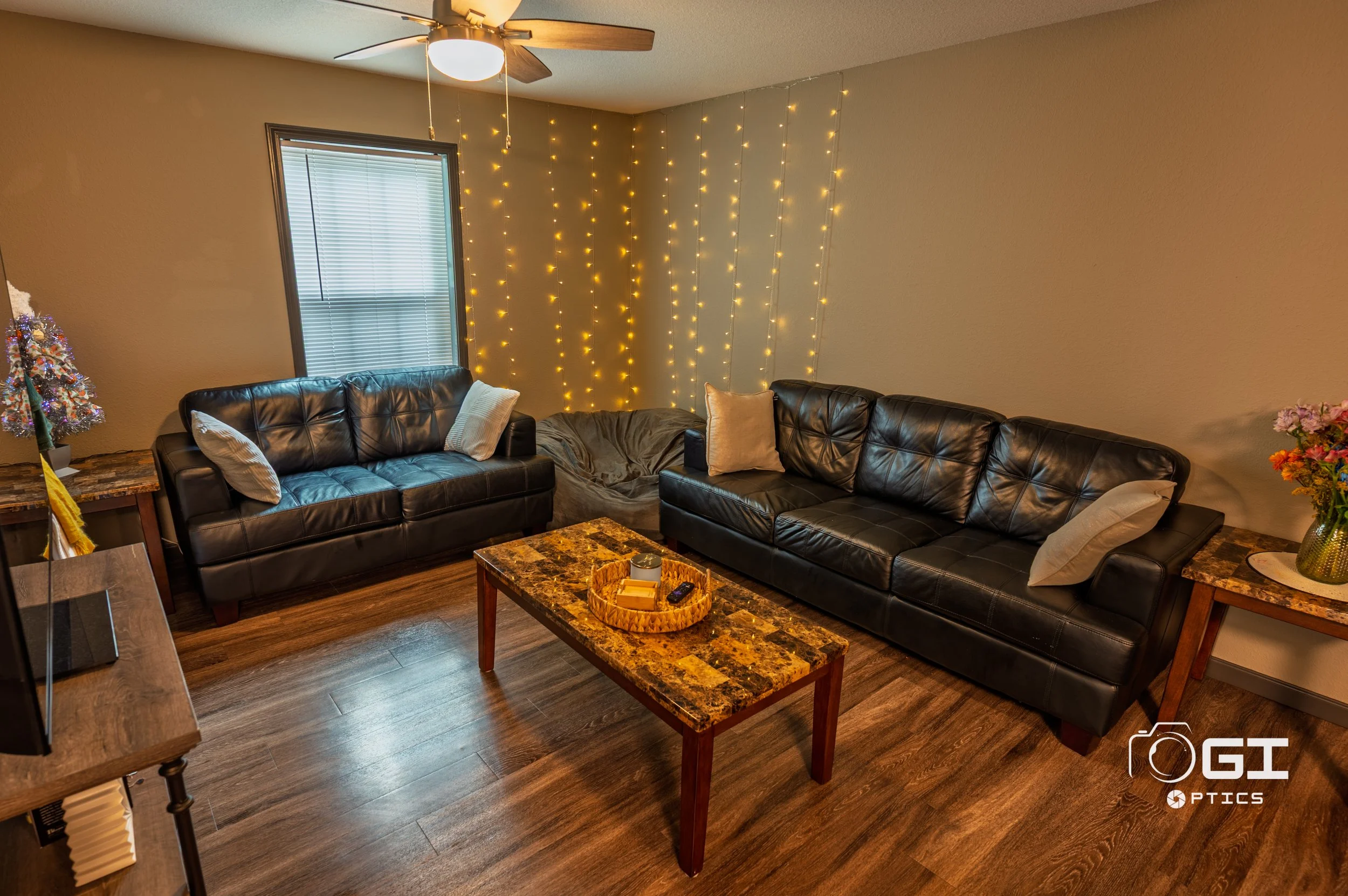Living room with two black leather sofas, a wooden coffee table with a tray holding books and a remote, a window with blinds, string lights on the wall, a ceiling fan, a small decorated Christmas tree, and side tables with vases of flowers.