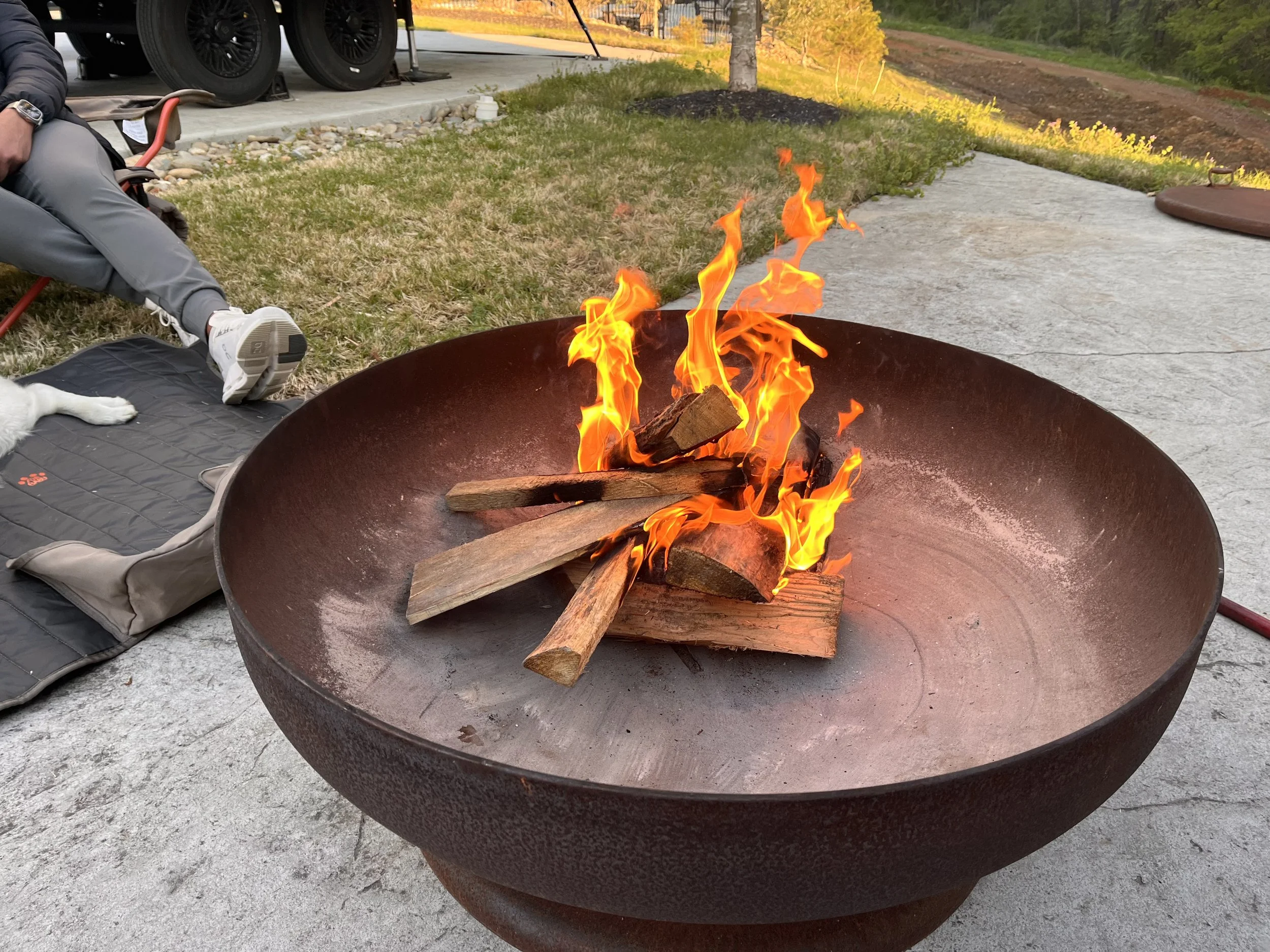 A fire pit with burning logs outside on a patio, with a person sitting nearby and their dog lying on a mat.