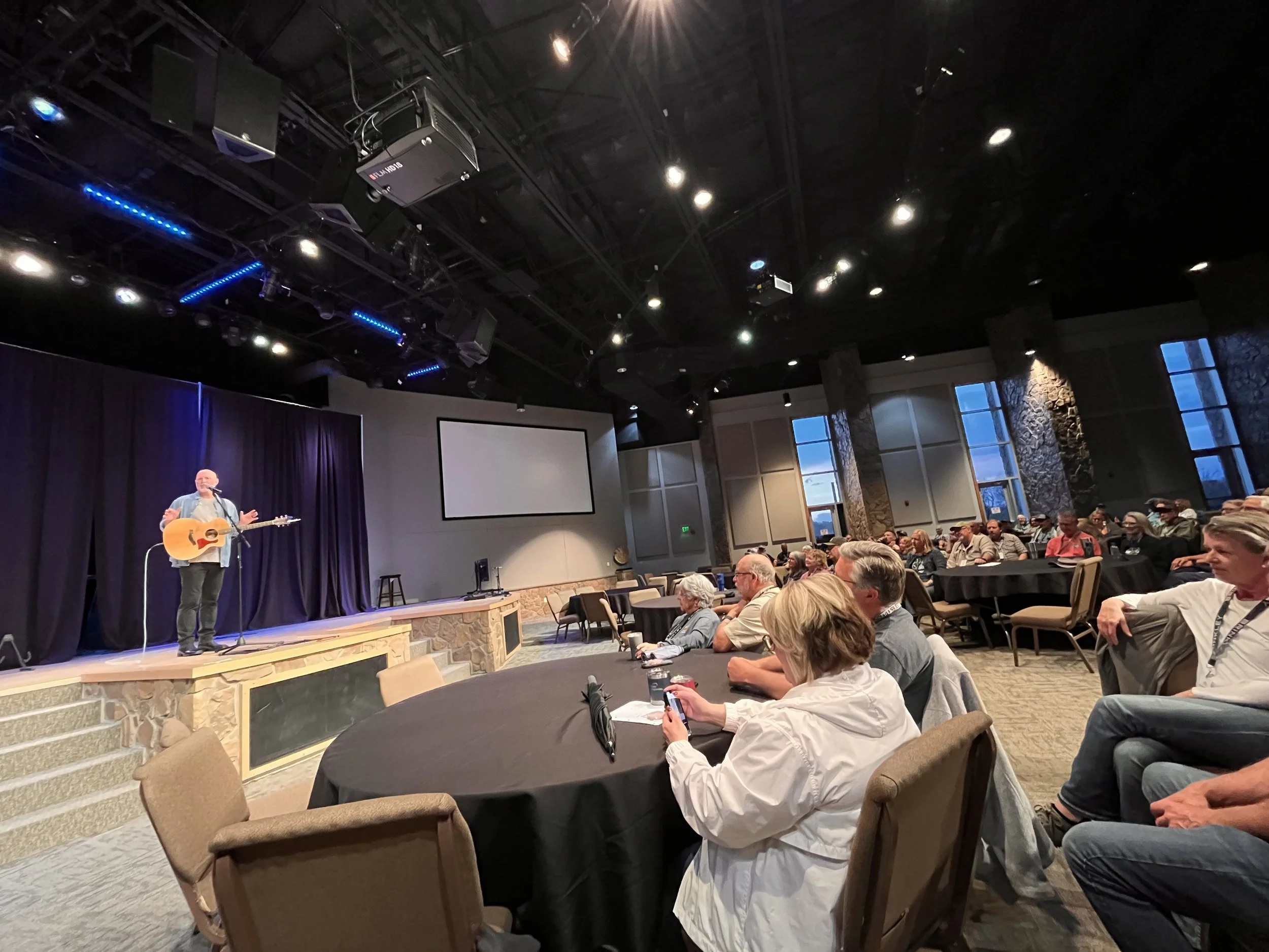 A man is performing a musical show on stage with a guitar, and an audience is seated at round tables attentively watching him.