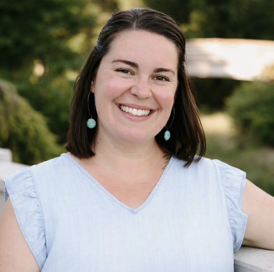 A woman with dark hair and earrings, smiling and leaning on a white railing outdoors with green trees in the background.