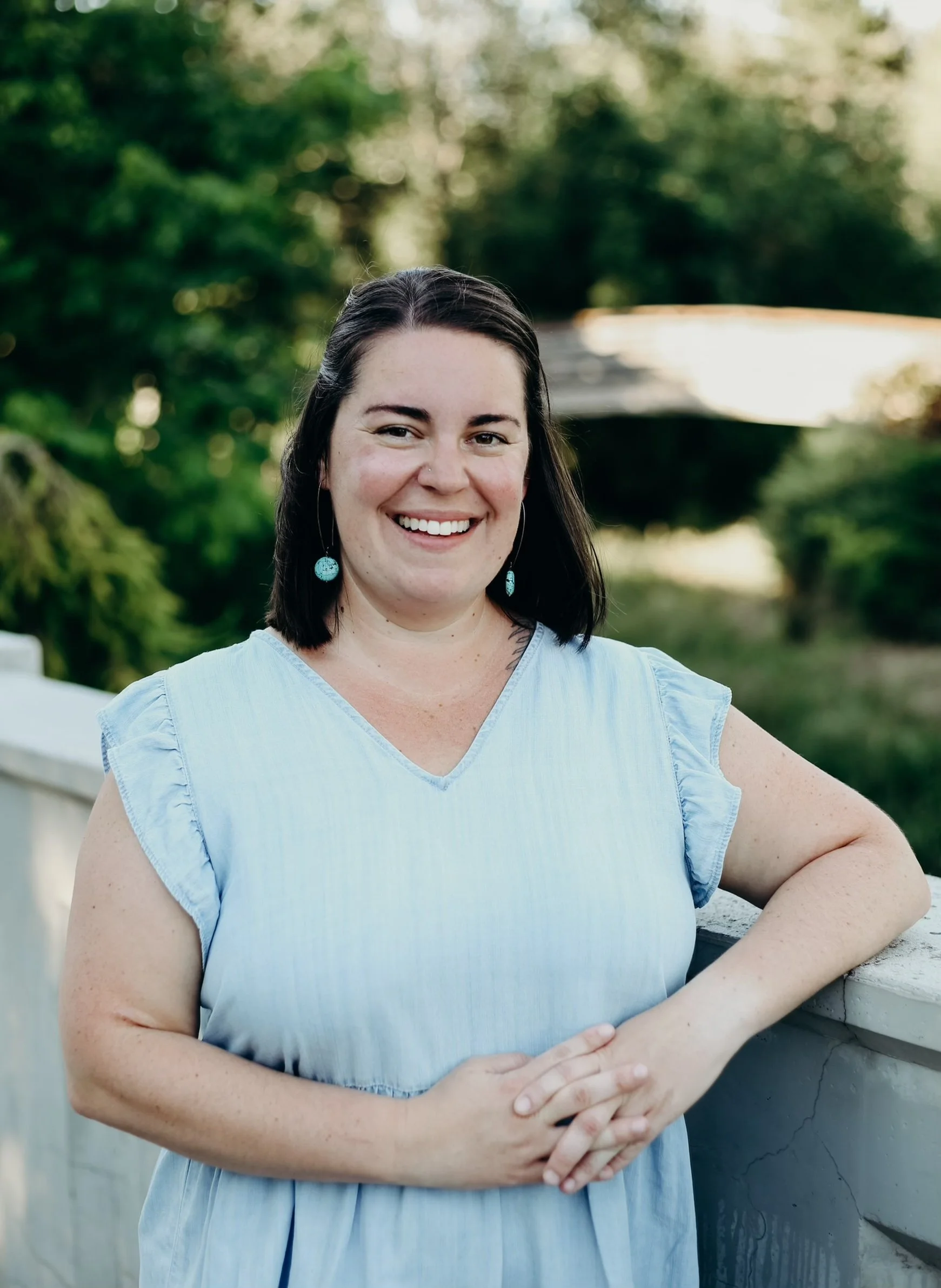 A woman with shoulder-length dark hair smiling, wearing a light blue sleeveless dress with ruffled sleeves and turquoise earrings, standing outdoors with greenery in the background.