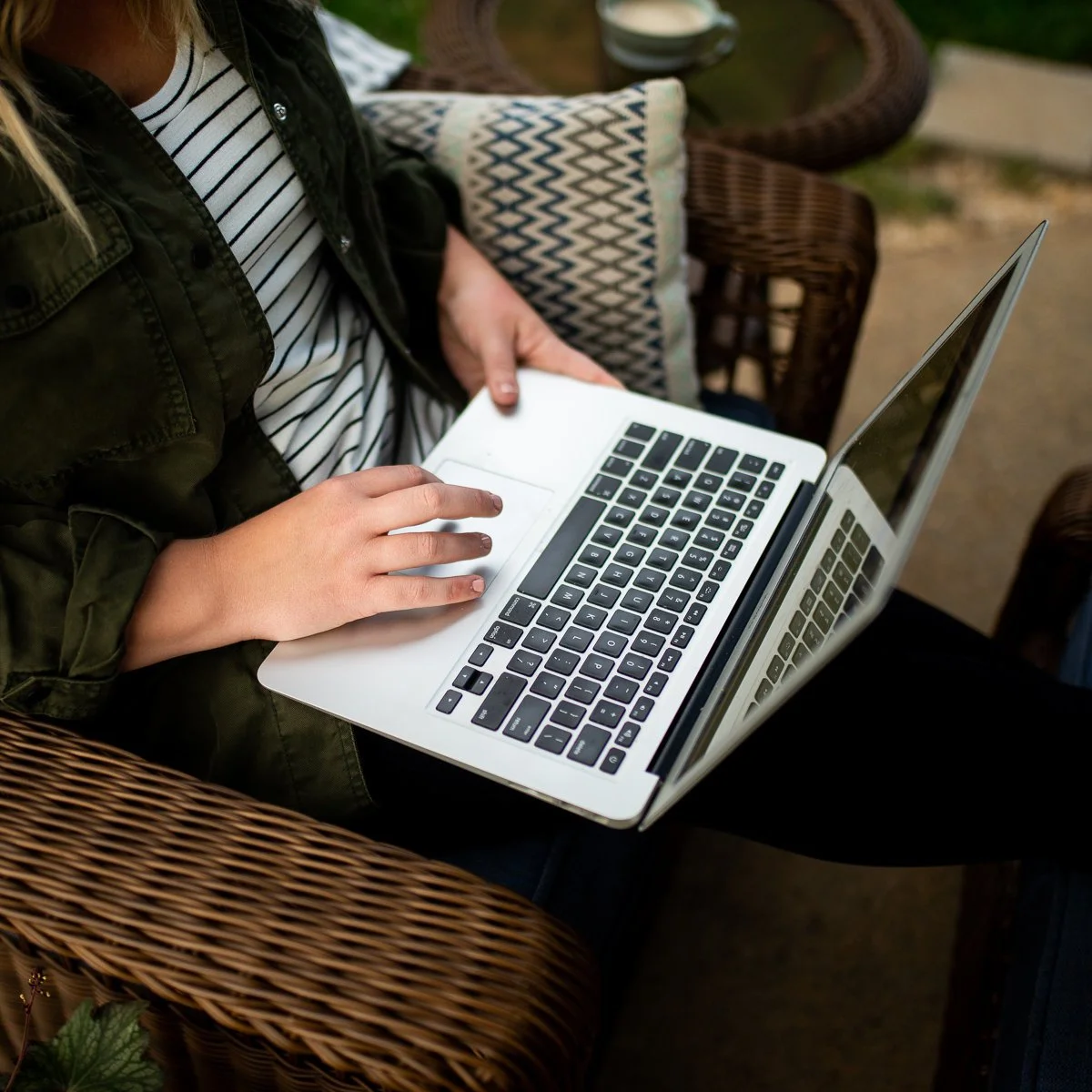 Person using a laptop while sitting on a wicker chair outdoors, with a cushion and table nearby.