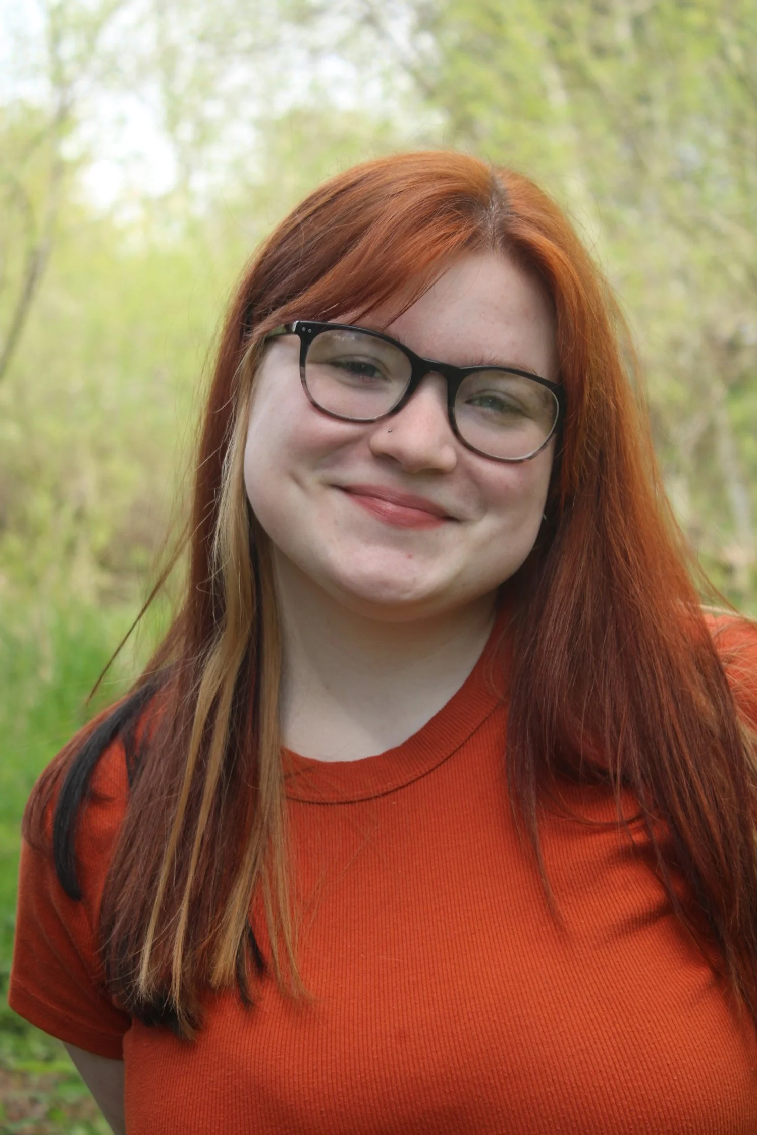 Portrait of a young woman with red hair, glasses, and a nose piercing, smiling outdoors in a green, wooded area.