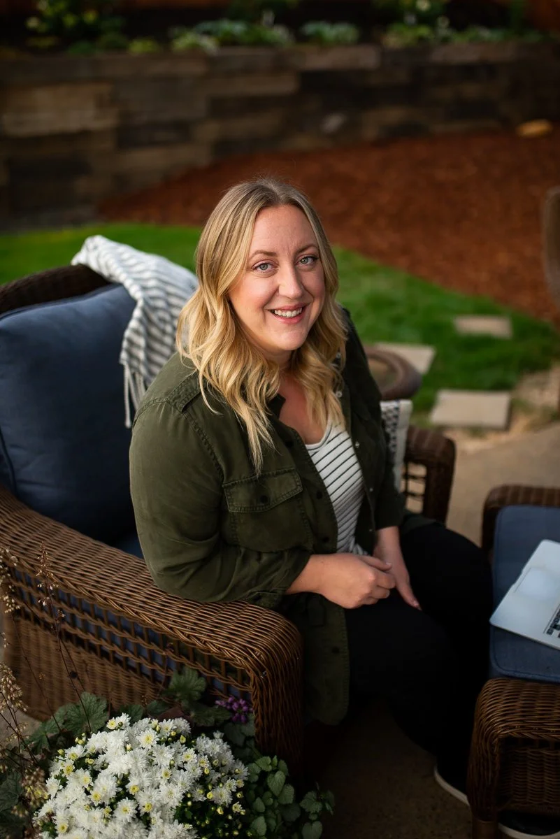 A woman with blonde wavy hair smiling while sitting outside on a patio chair with a laptop on her lap, surrounded by flowers and greenery.