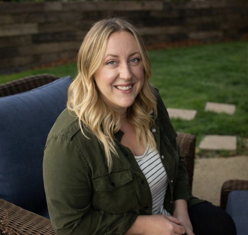 A woman with blonde hair smiling, sitting on a wicker chair outside. She is wearing a green jacket over a striped shirt, with a laptop on her lap. There are flowers in the foreground and a wooden fence in the background.