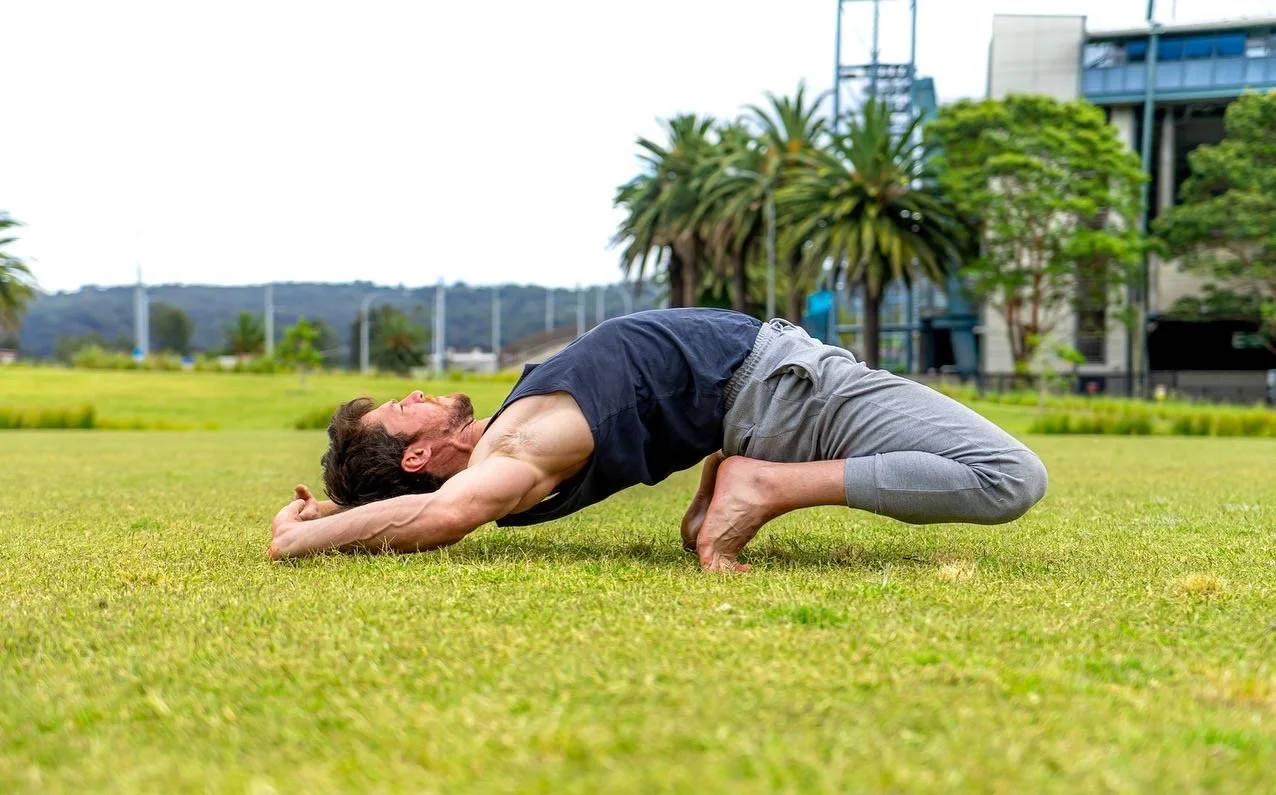 Outdoor movement practice in a grassy field, exploring hip mobility and grounded posture with an urban backdrop.