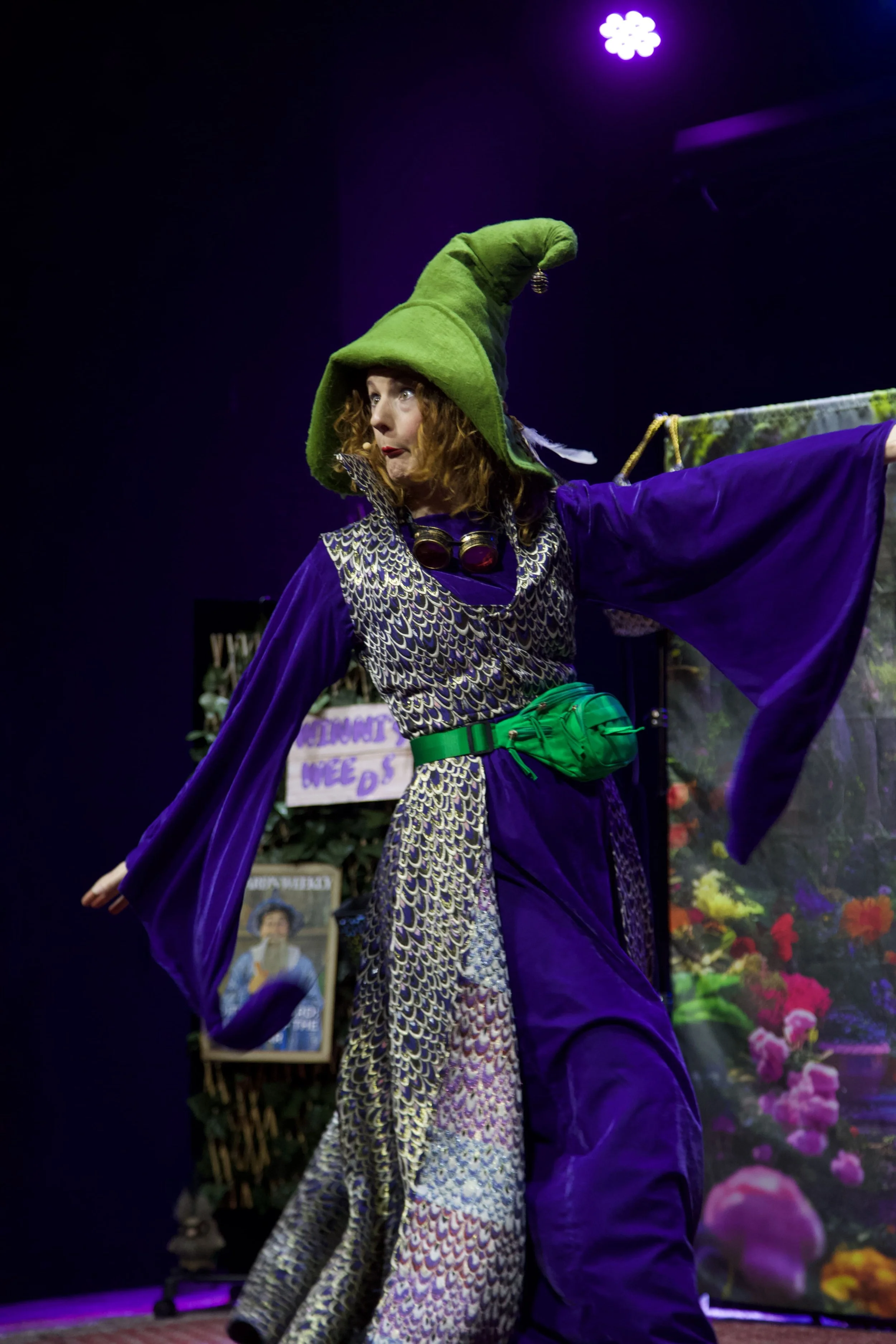 Woman dressed as a wizard with a green hat and purple cloak performing on stage with a colorful background.
