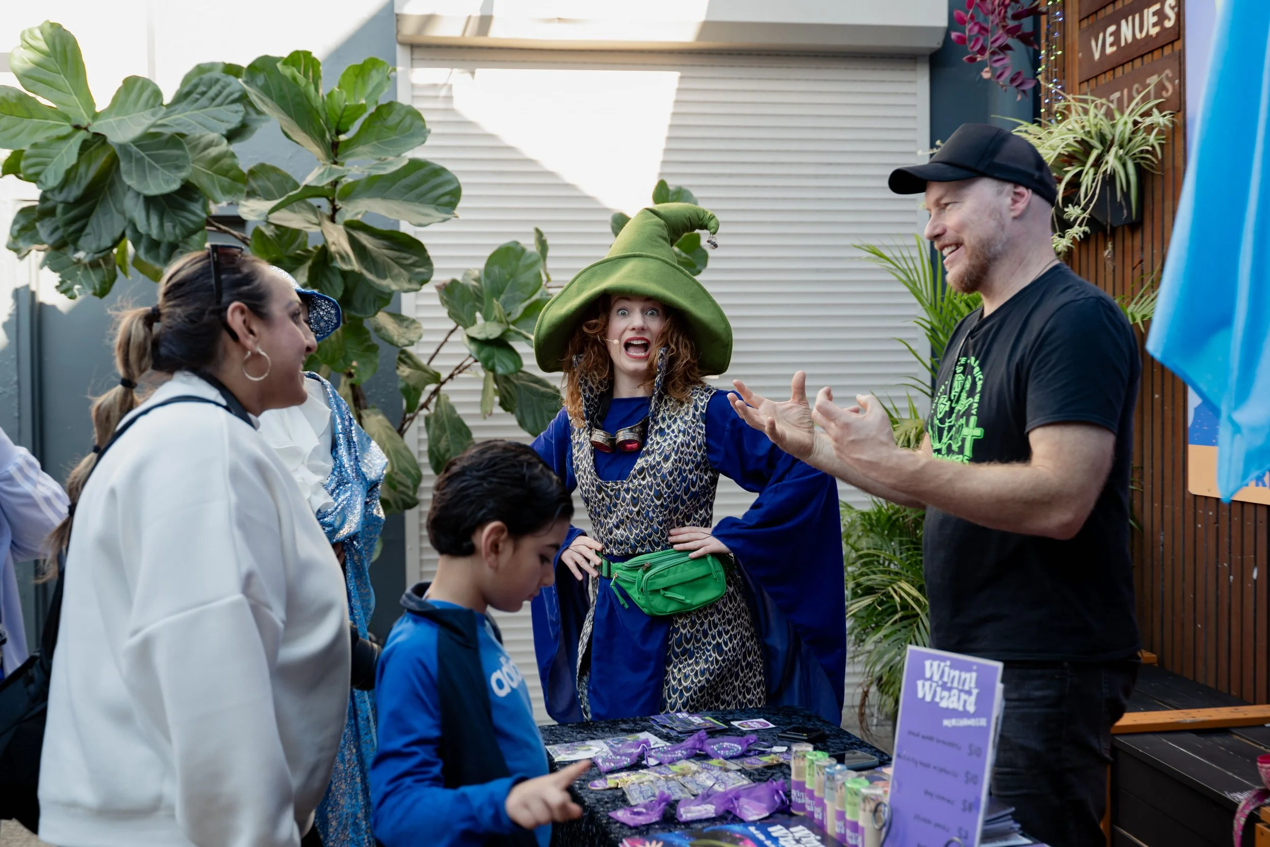 A woman dressed as a wizard with a large green hat and a colorful costume, speaking animatedly to a group at an outdoor event. Several people are gathered around a booth with a sign that reads 'Winning Wizar'. Large plants and decorative banners are 