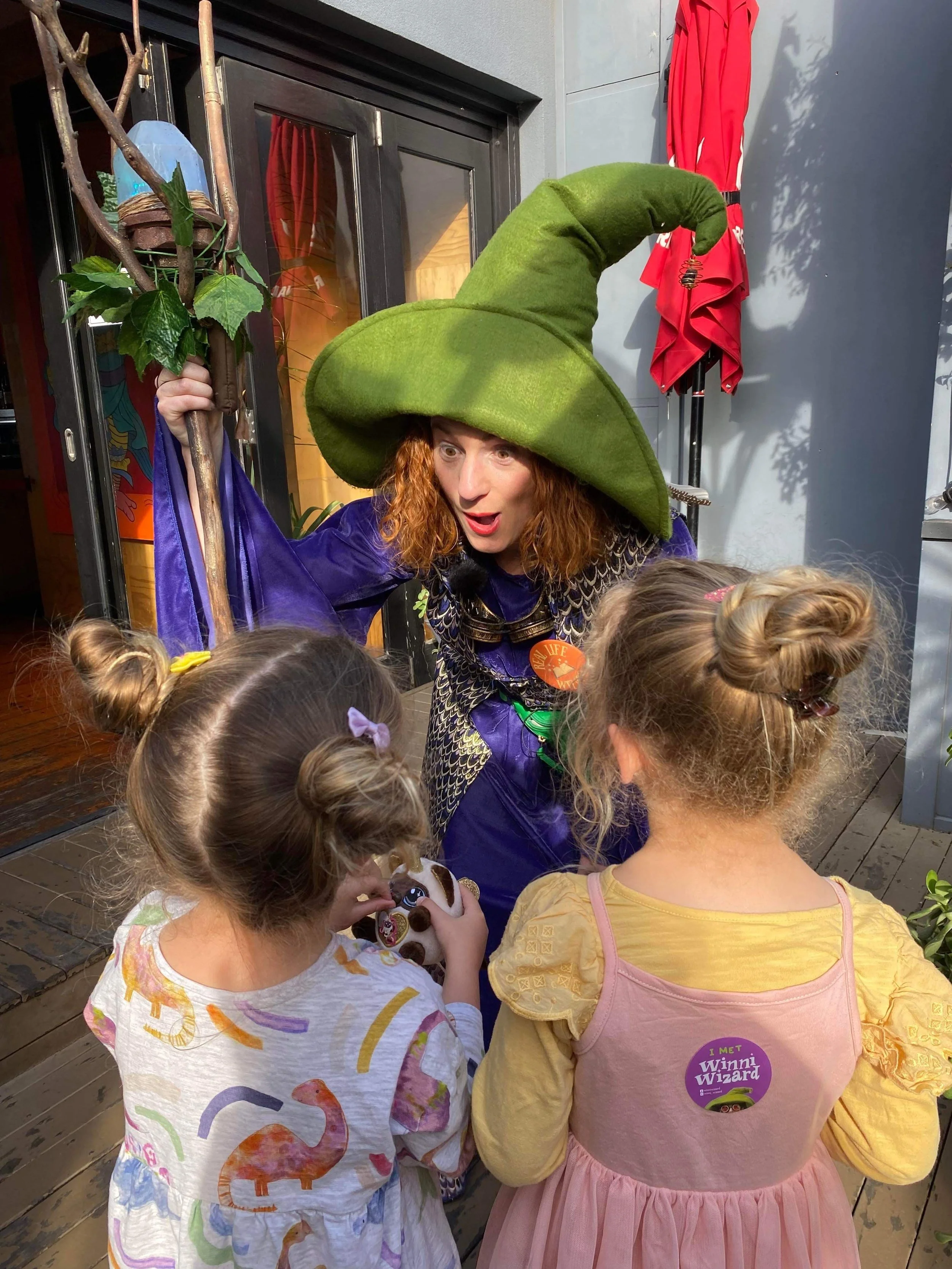 A woman dressed as a witch with a big green hat, purple cloak, and costume, talking to two young girls with their backs to the camera. The woman appears to be entertaining the children.