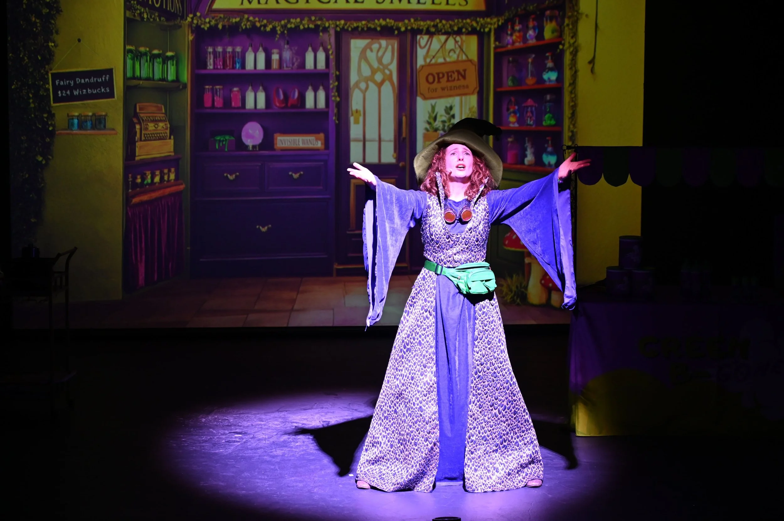 A woman dressed as a wizard character performing on stage. She is wearing a wide-brimmed hat, a leopard print top under a purple cloak, a long patterned skirt, a green sash with a green pouch, and spooky goggles around her neck. She stands with arms 