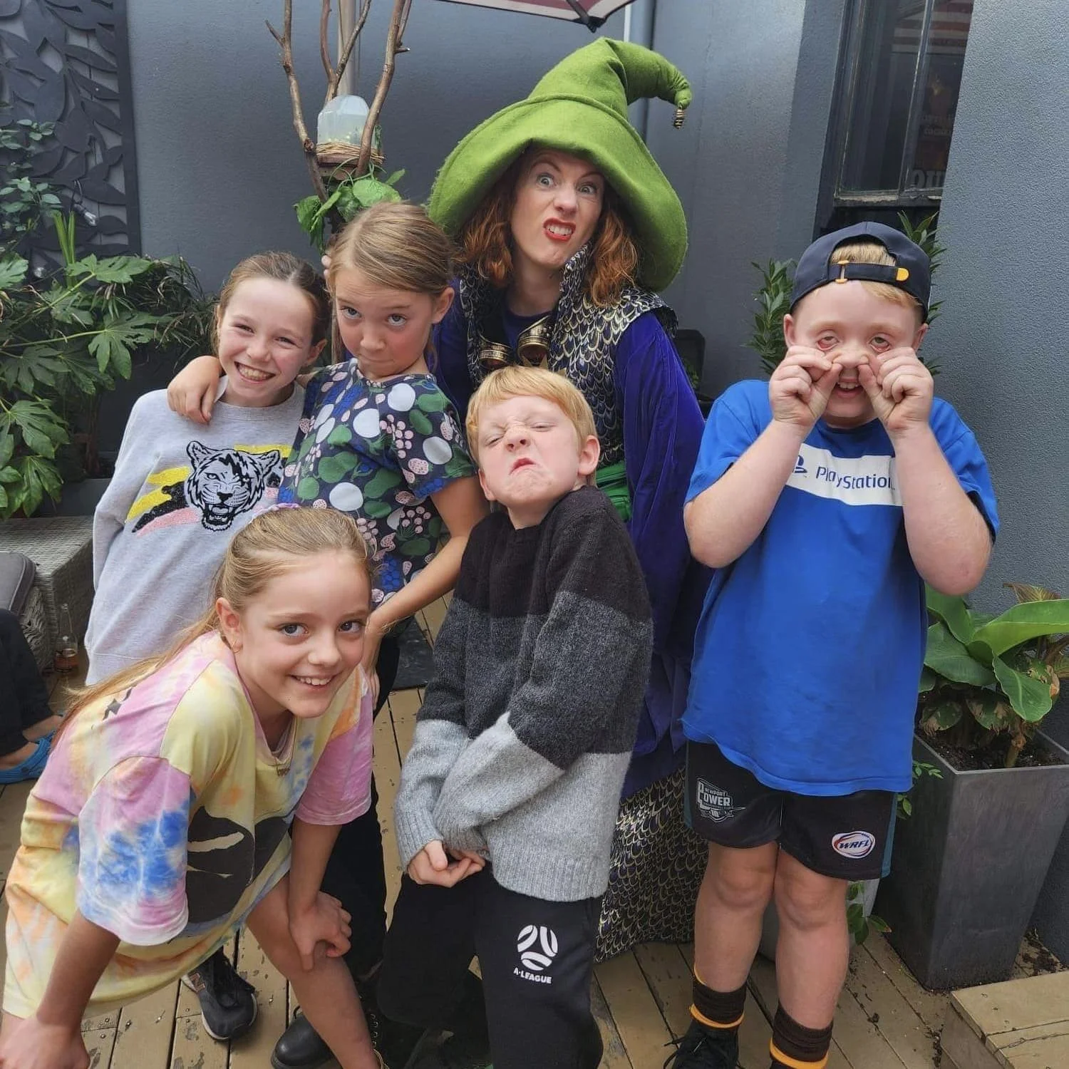 Group of six children with an adult dressed as a witch, making funny faces and poses in an outdoor setting with plants and gray walls.