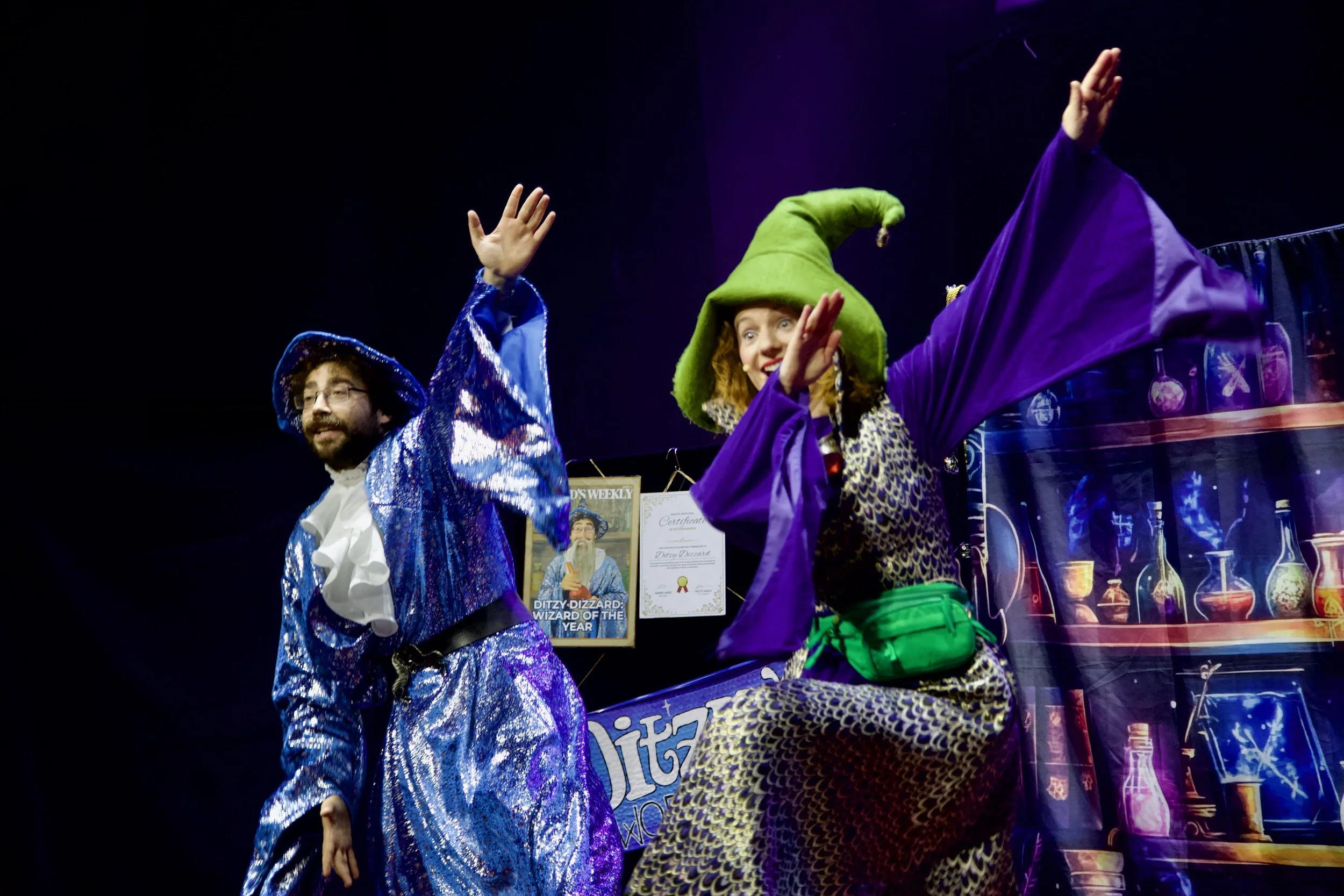 Two performers in colorful costumes on stage, one dressed as a wizard with a sparkly blue robe and hat, the other as a witch with a green hat, purple robes, and patterned pants, engaging in a lively act.