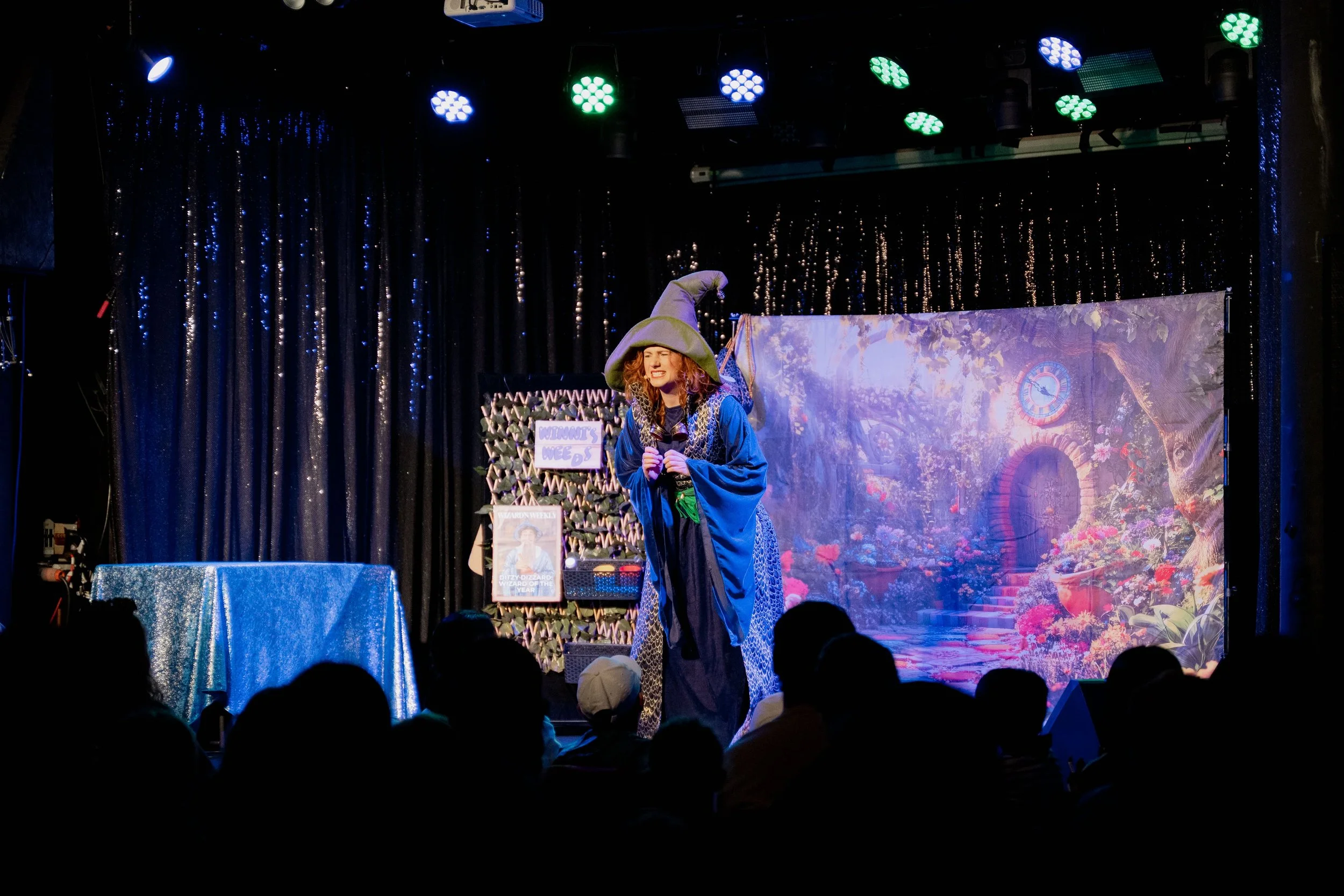 A woman dressed as a wizard on stage during a performance, wearing a large green wizard hat and a long blue robe with patterned accents. The stage has a backdrop of a enchanted garden scene with flowers, steps, and a clock. Audience members are visib