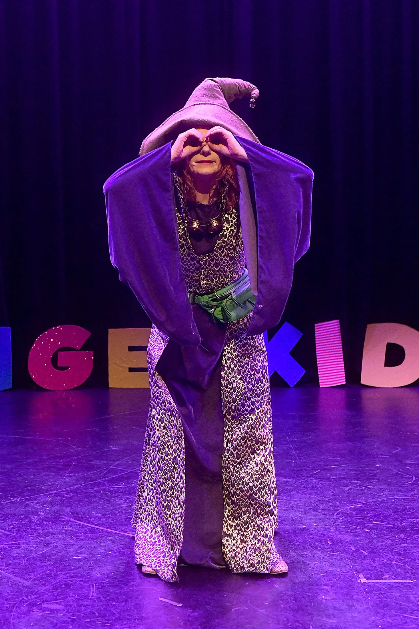 A person dressed in a purple and gold costume with a large, floppy hat, standing on a stage with colorful letters behind them. The person is making a heart shape with their hands around their eyes.