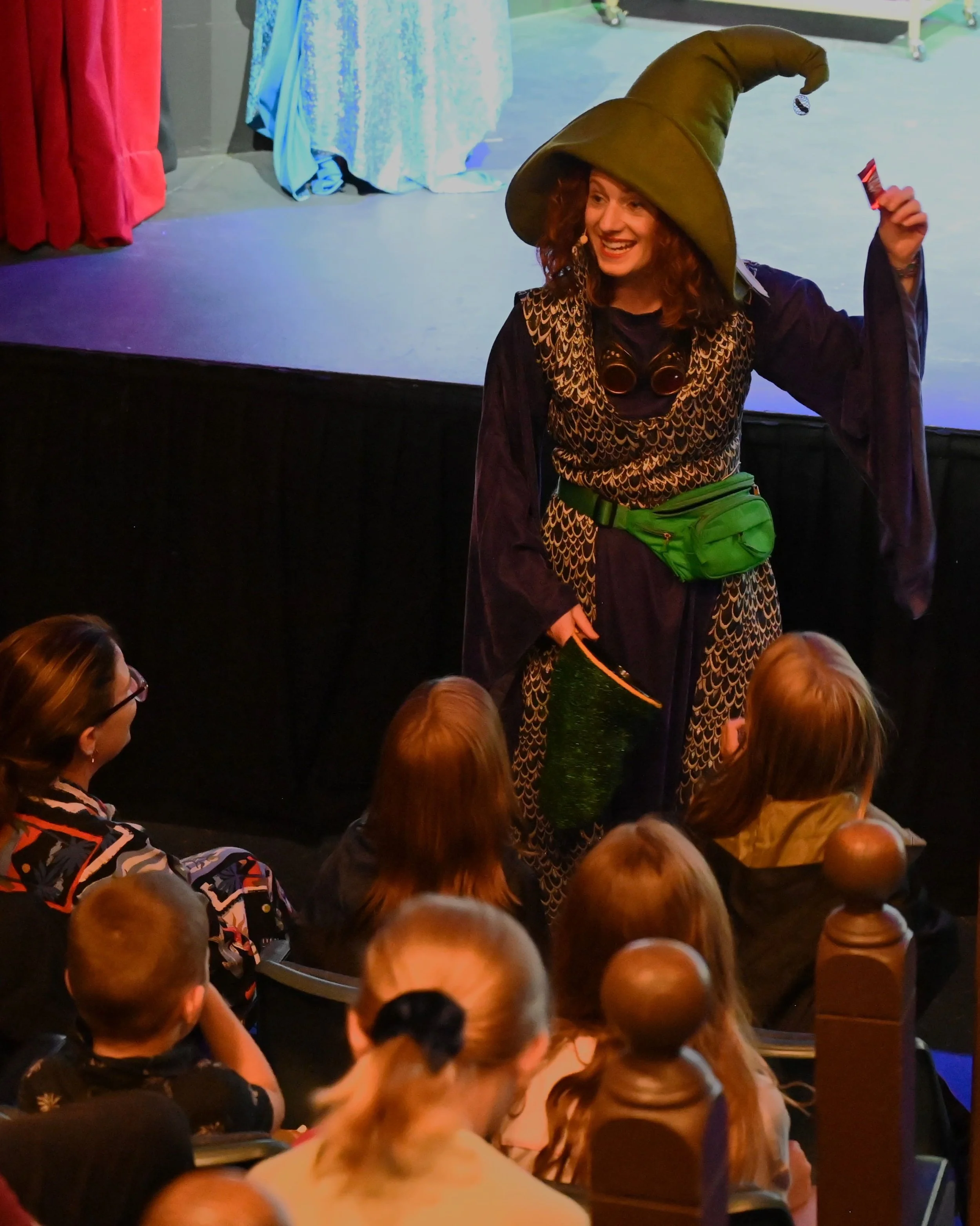 A woman dressed as a wizard with a large green hat, dark cloak, and a green pouch on her waist entertains children in an audience during a show.