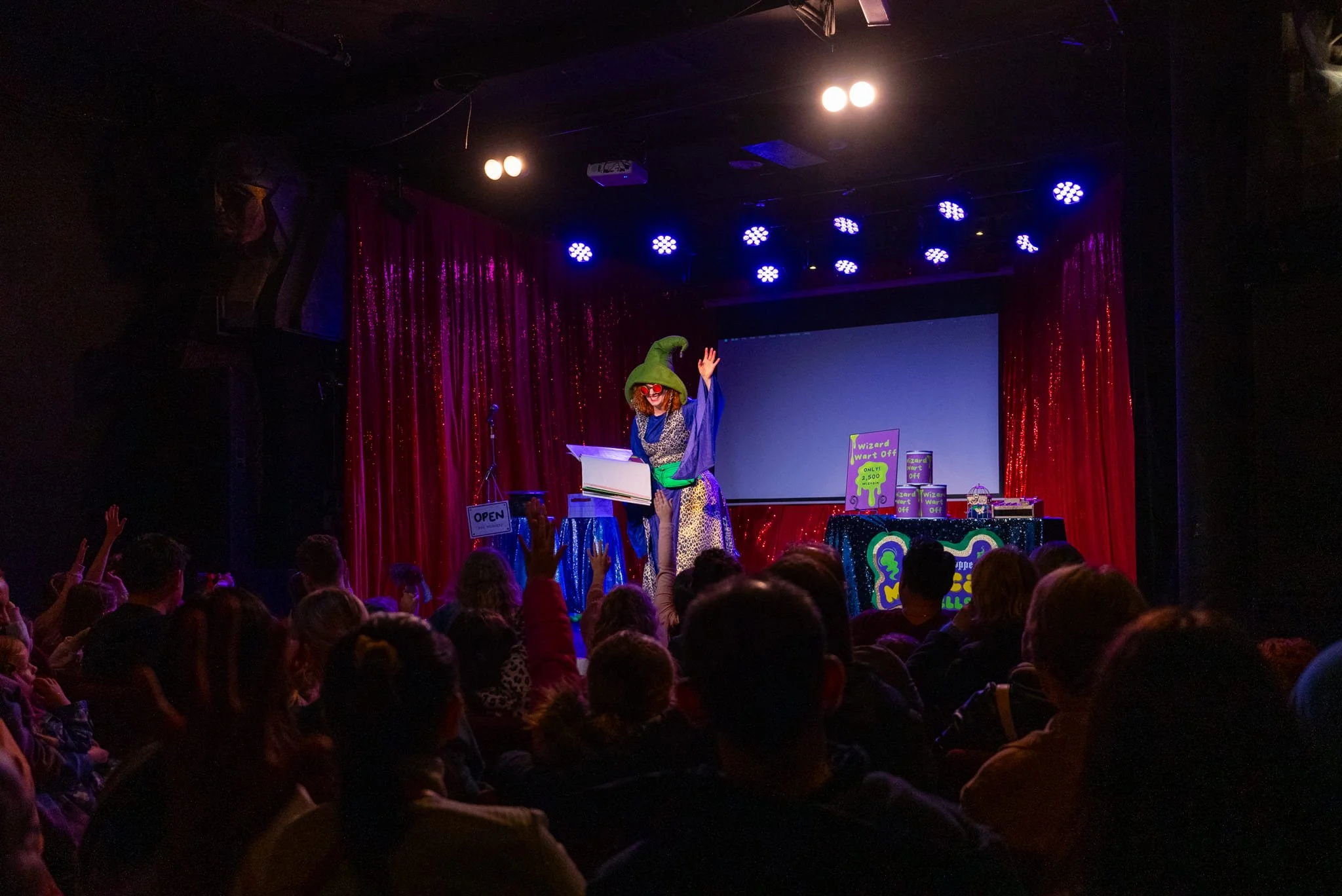 Children in an audience watching a magician on stage perform a magic trick, with some children raising their hands. The magician is dressed as a wizard with a green hat, glasses, and a colorful outfit, standing in front of a red curtain with blue and