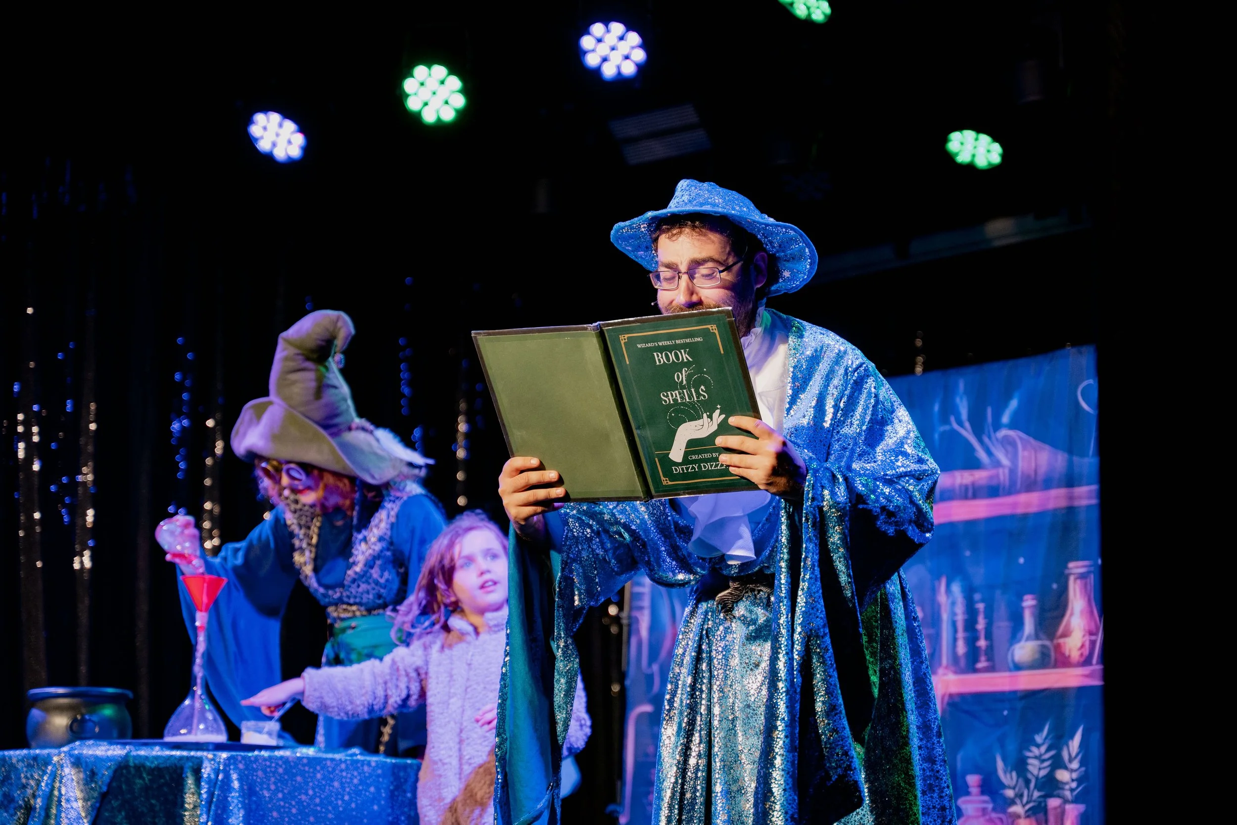 A man dressed as a wizard wearing a blue sparkly robe and matching pointed hat reads a book titled 'Book of Spells' on stage with children in costume, including a girl and a witch with a large hat, against a black backdrop with colorful stage lights.
