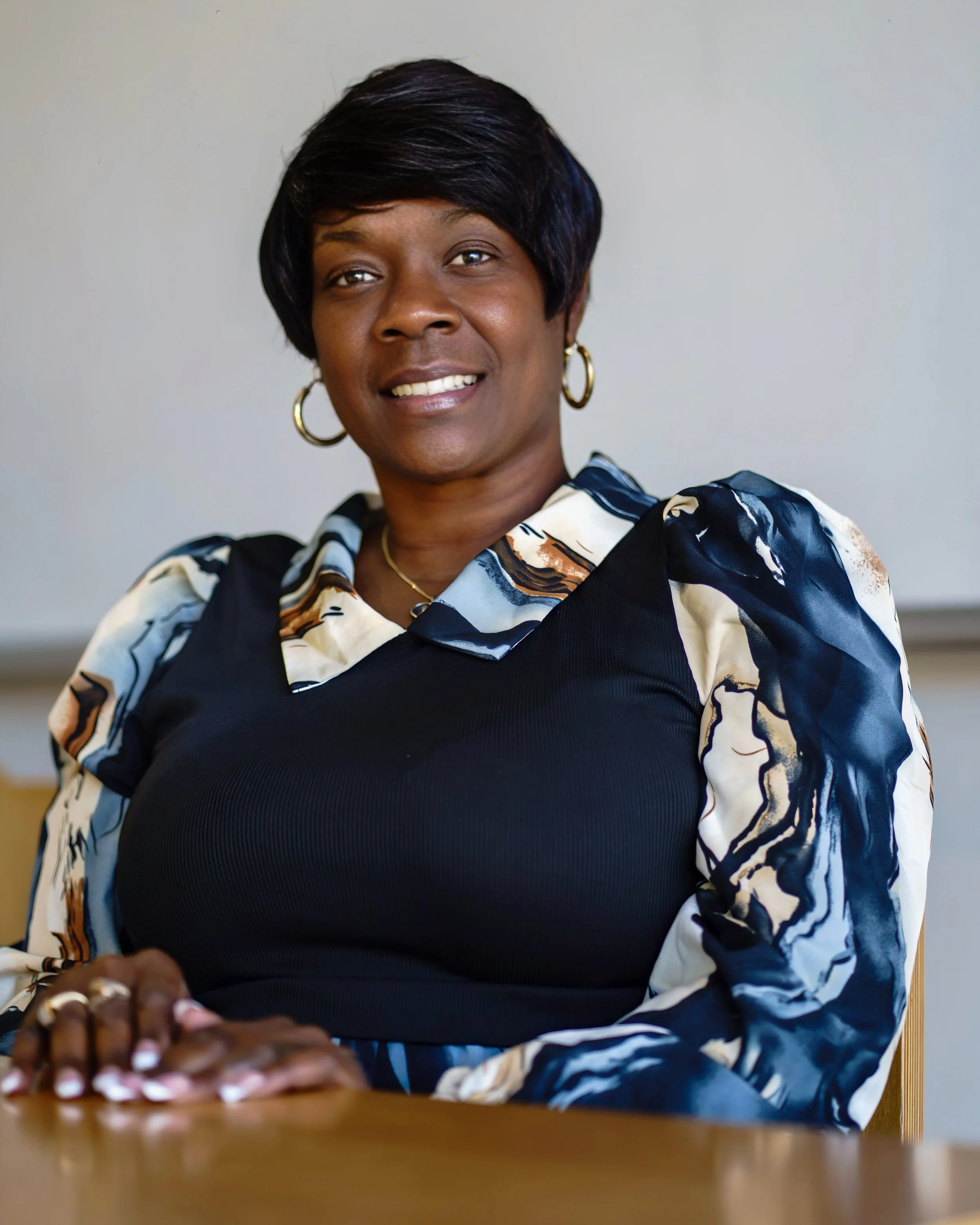 A woman with short black hair, wearing gold hoop earrings, a gold necklace, and a black top with patterned sleeves, sitting at a desk and smiling.