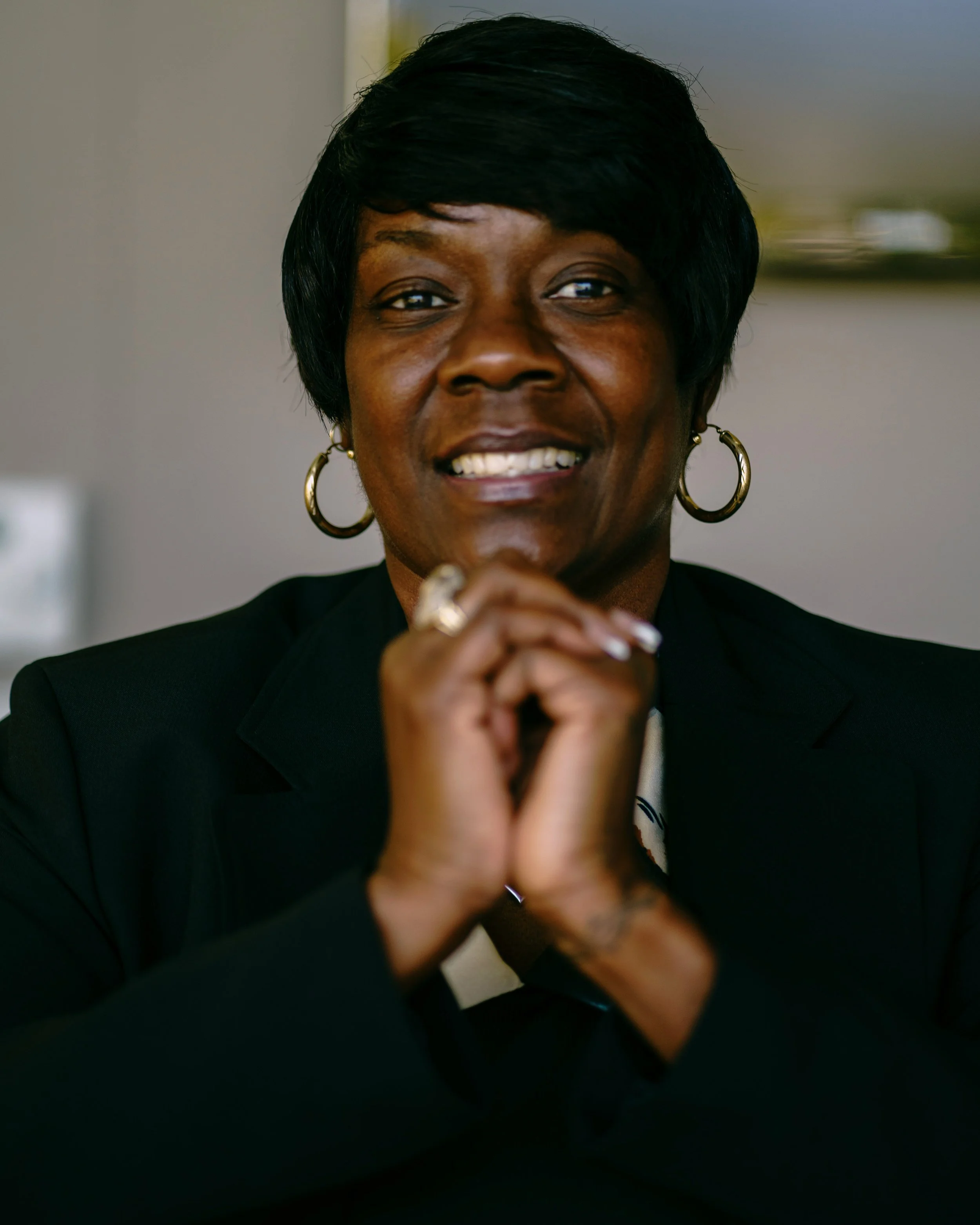 A smiling Black woman with short black hair, wearing hoop earrings, a black blazer, and a ring, holding her hands together near her face.