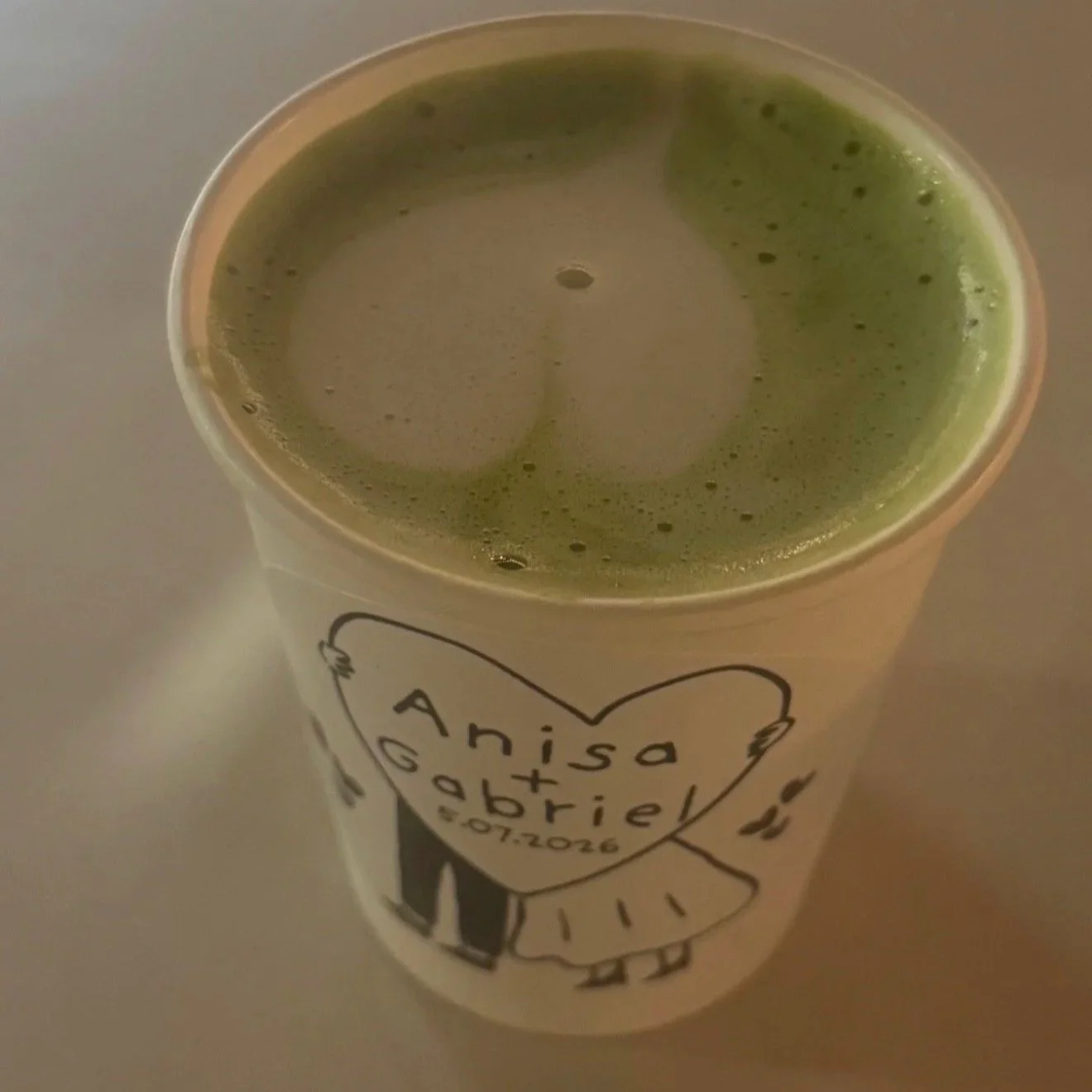 A cup of green smoothie with foam on top and a label with a drawing of a girl and the words 'Anisa, Gedied, 07.07.2016'.