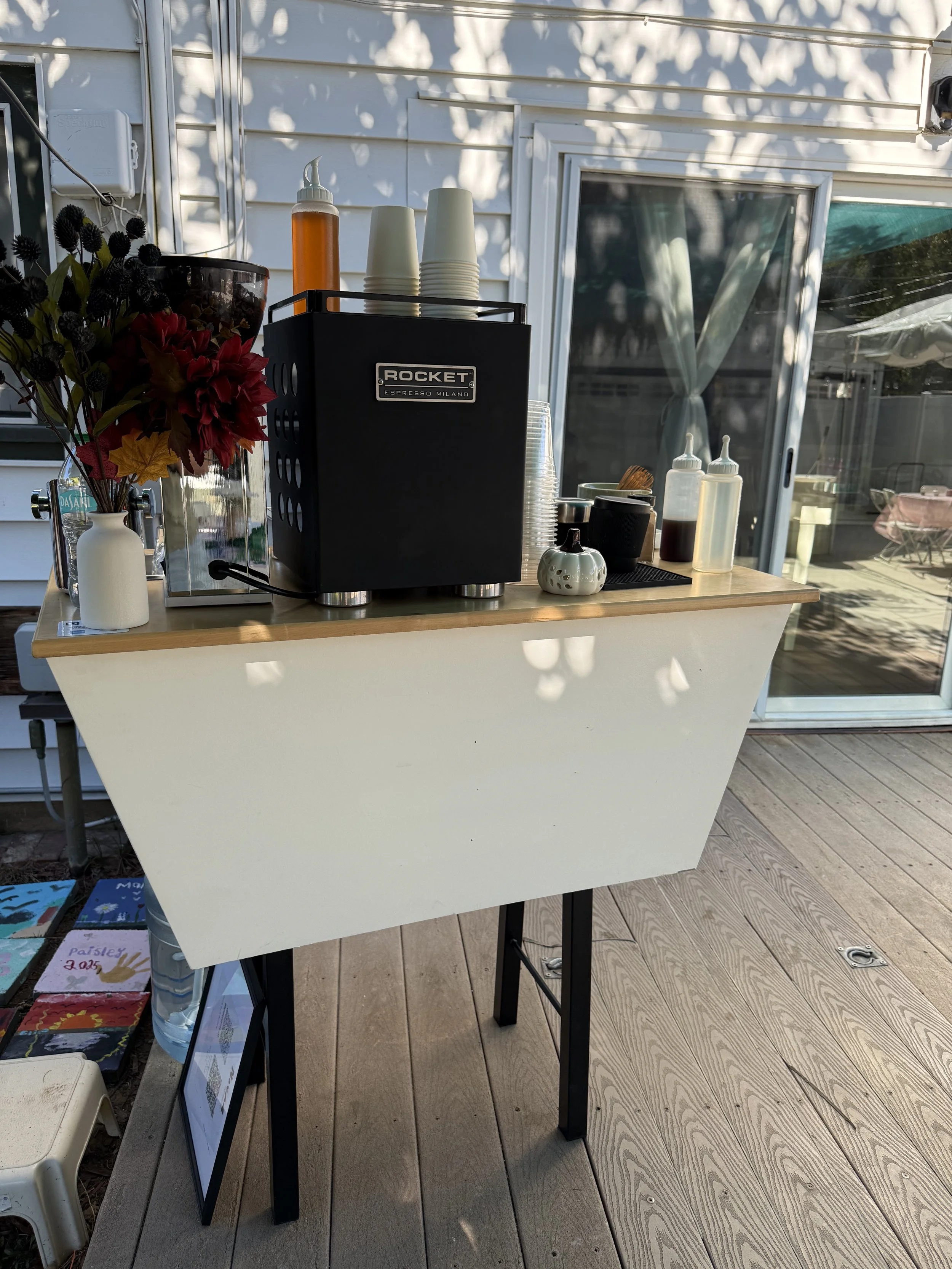 A outdoor espresso catering bar stand with a Rocket espresso machine on a wooden coffee cart, surrounded by cups, syrup bottles, and decorations, with a sliding glass door in the background.
