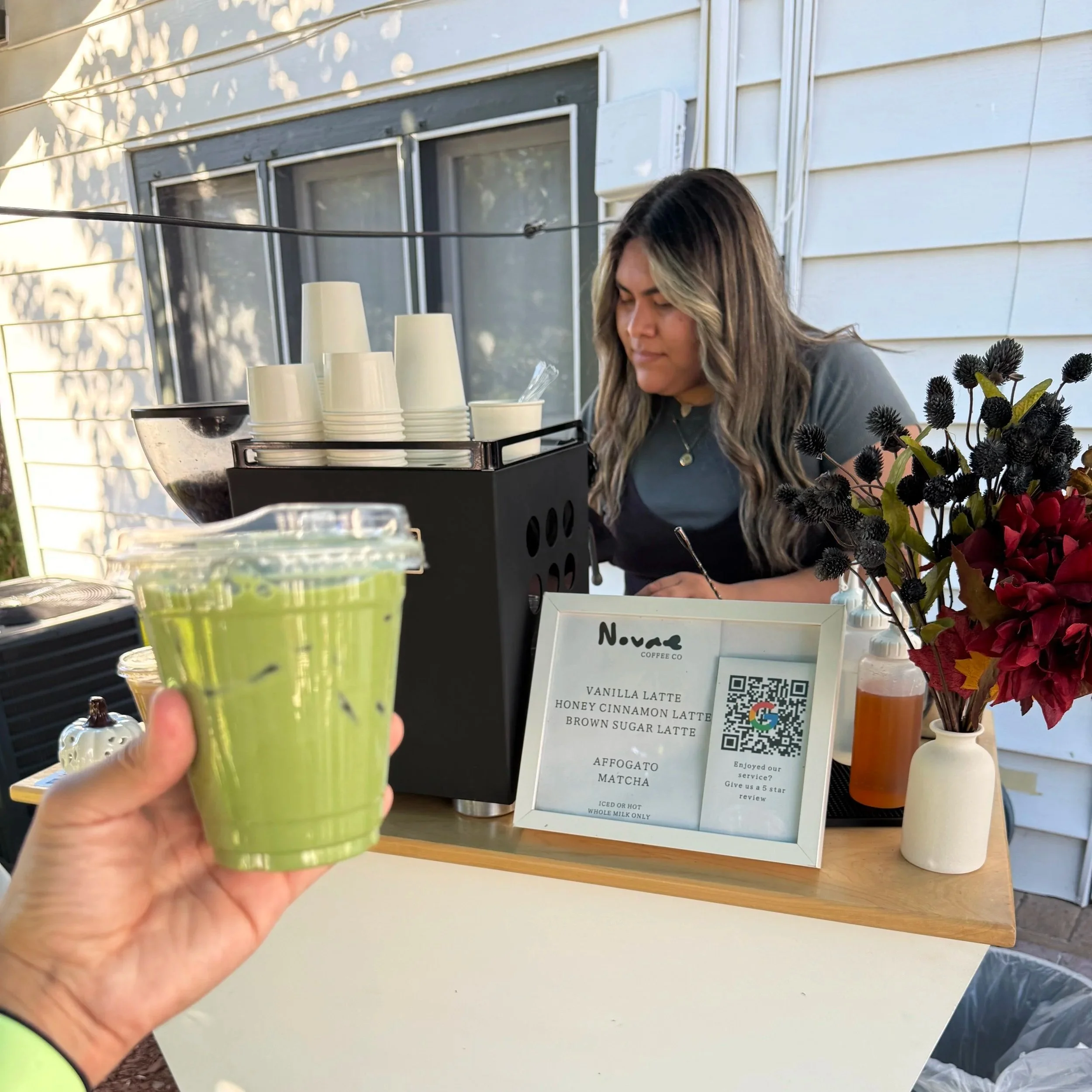 A person holding a bright green beverage in a plastic cup with a straw at an outdoor coffee stand. The background includes stacks of white cups, a menu sign listing different latte flavors, and a small vase of dark purple and red flowers on a wooden table.
