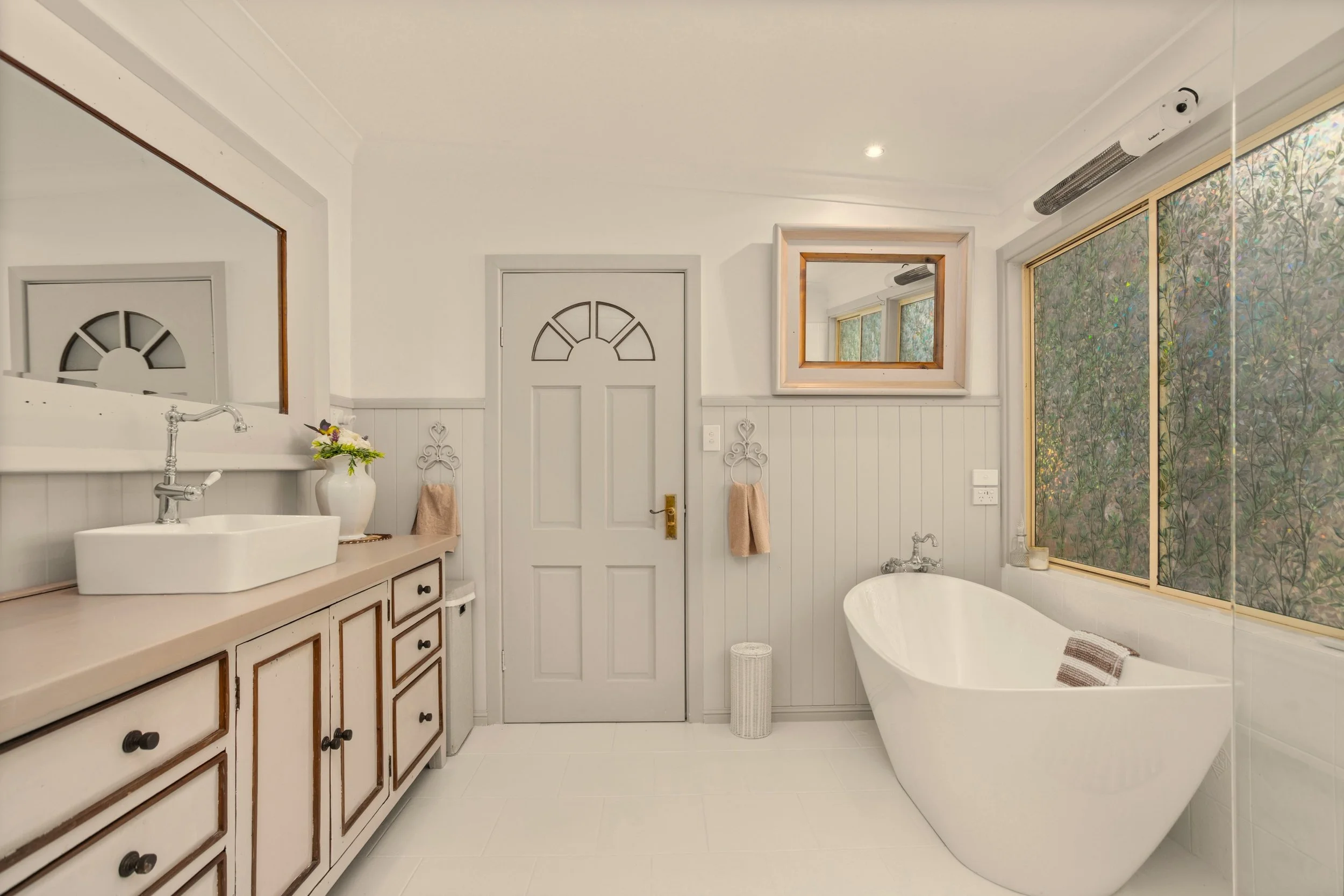 Bathroom with a modern white bathtub, a vanity with a vessel sink, mirror, wall decor, and large window with foliage outside