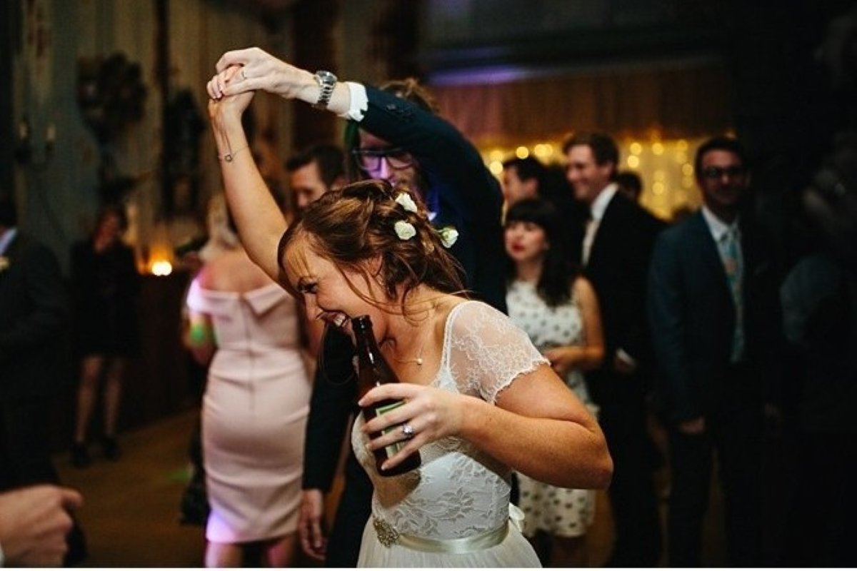 A woman in a white lace dress dancing at a party, holding a bottle and smiling with her eyes closed, while a man behind her twirls her around. People in formal attire are seen in the background at a celebration.