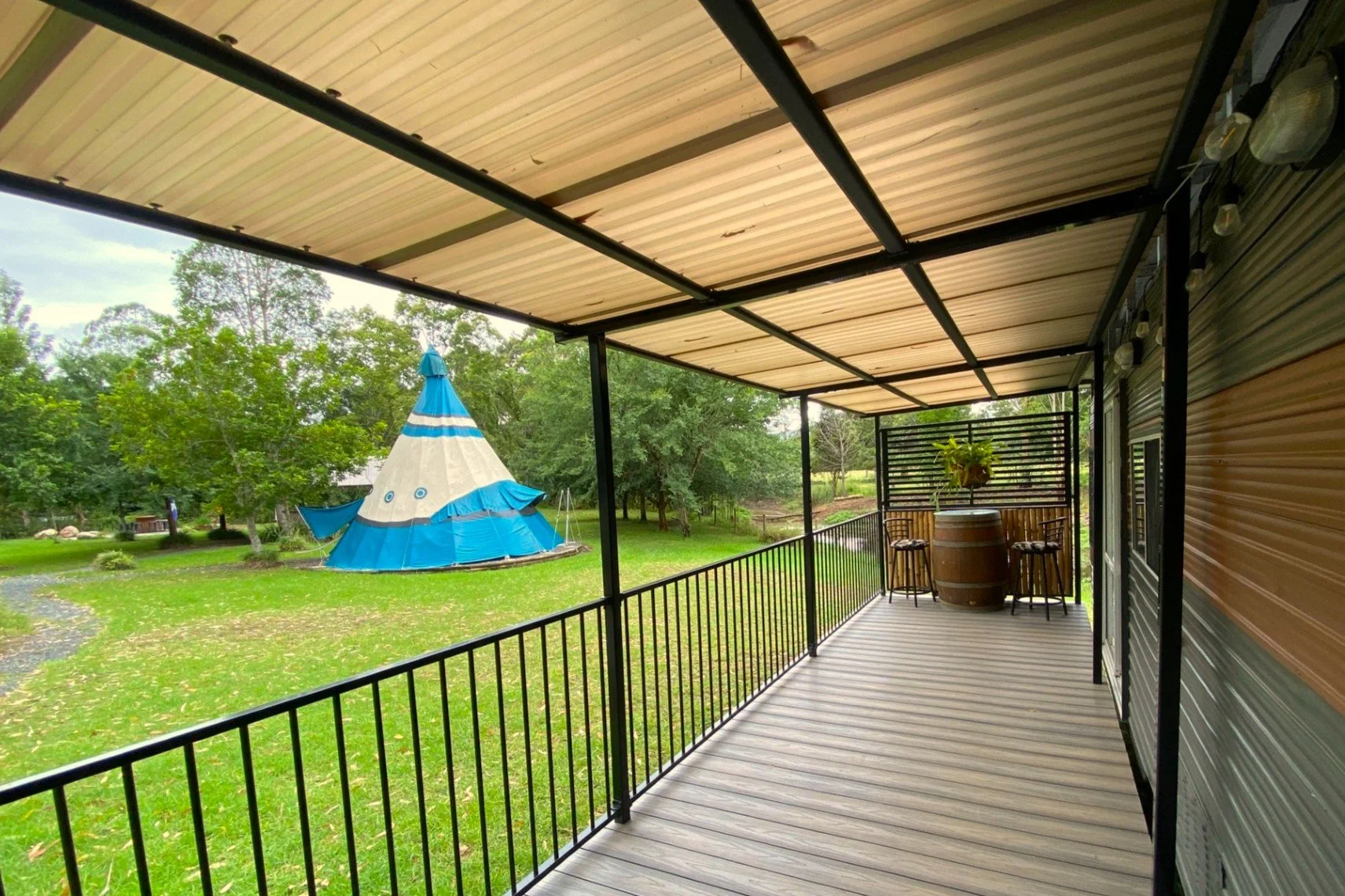 View from a covered porch with a black metal railing, overlooking a grassy yard with trees and a blue and white teepee tent.