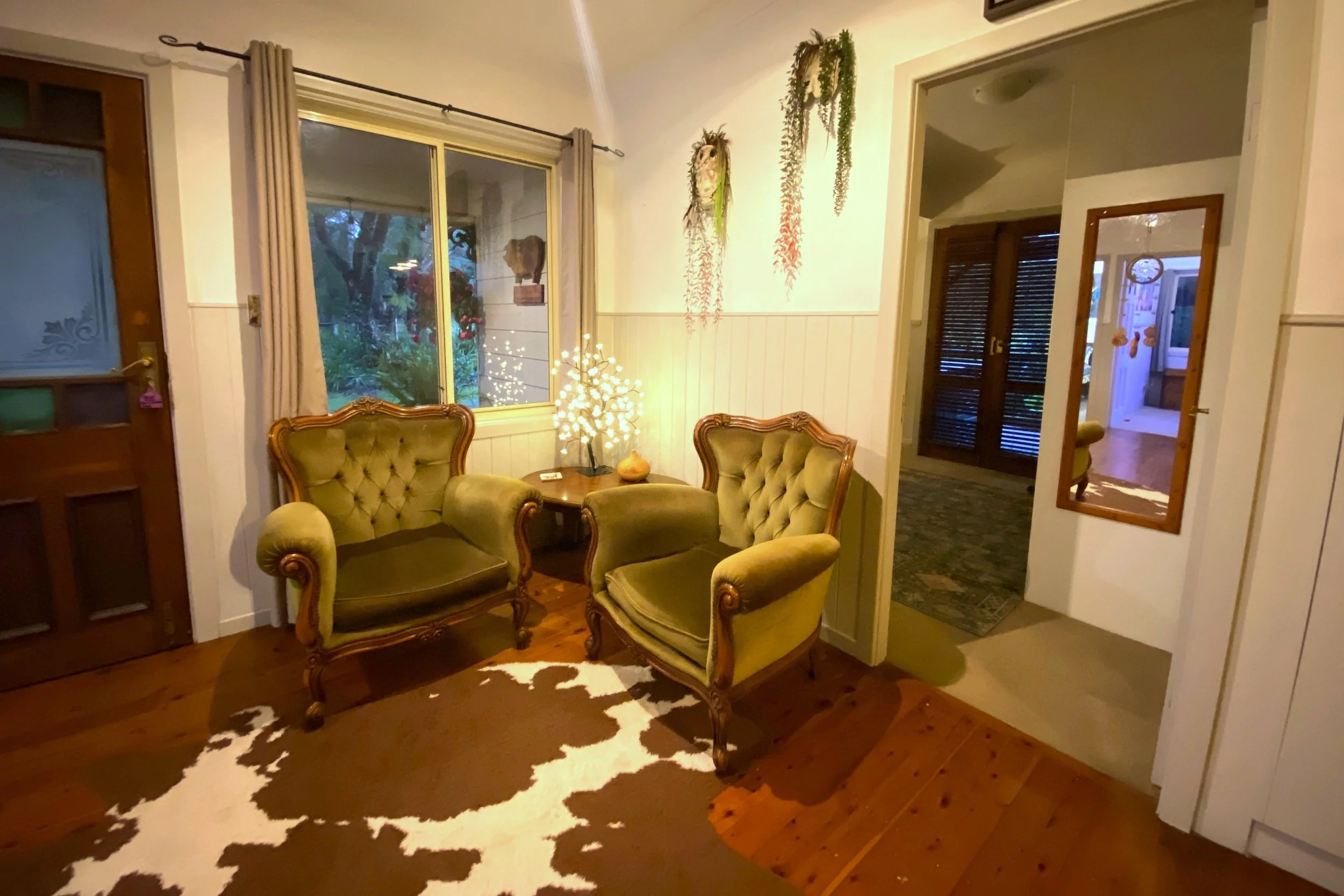 Living room with two vintage armchairs, a side table with a decorative bowl and illuminated tree, window with curtains, mirror on wall, hanging plants, and a cowhide rug on wooden floor.