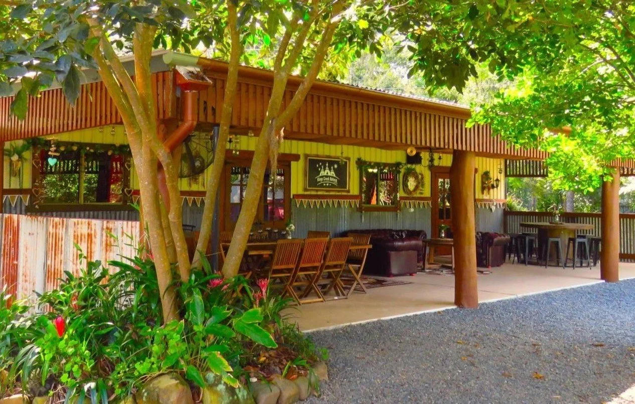 Outdoor patio area of a rustic restaurant or cafe with wooden furniture, some chairs around a table, bar stools, and lounge chairs. Surrounded by trees and plants, creating a cozy, inviting ambiance.