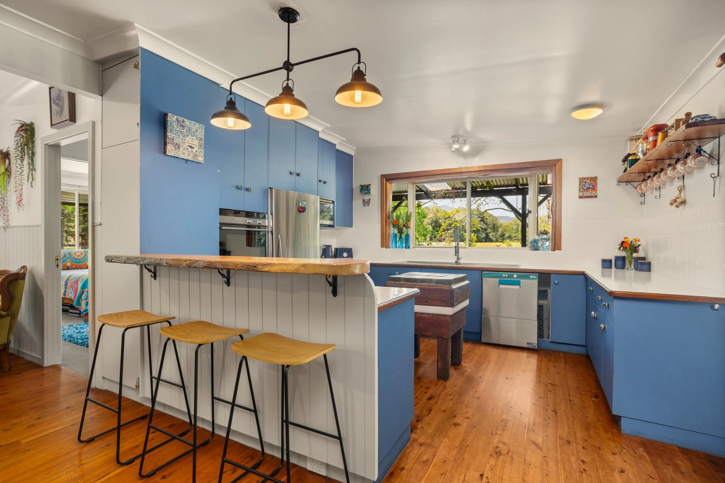 Kitchen with blue cabinets, wooden countertop, and three barstools, large window showing greenery outside, and open shelf with cups and jars.