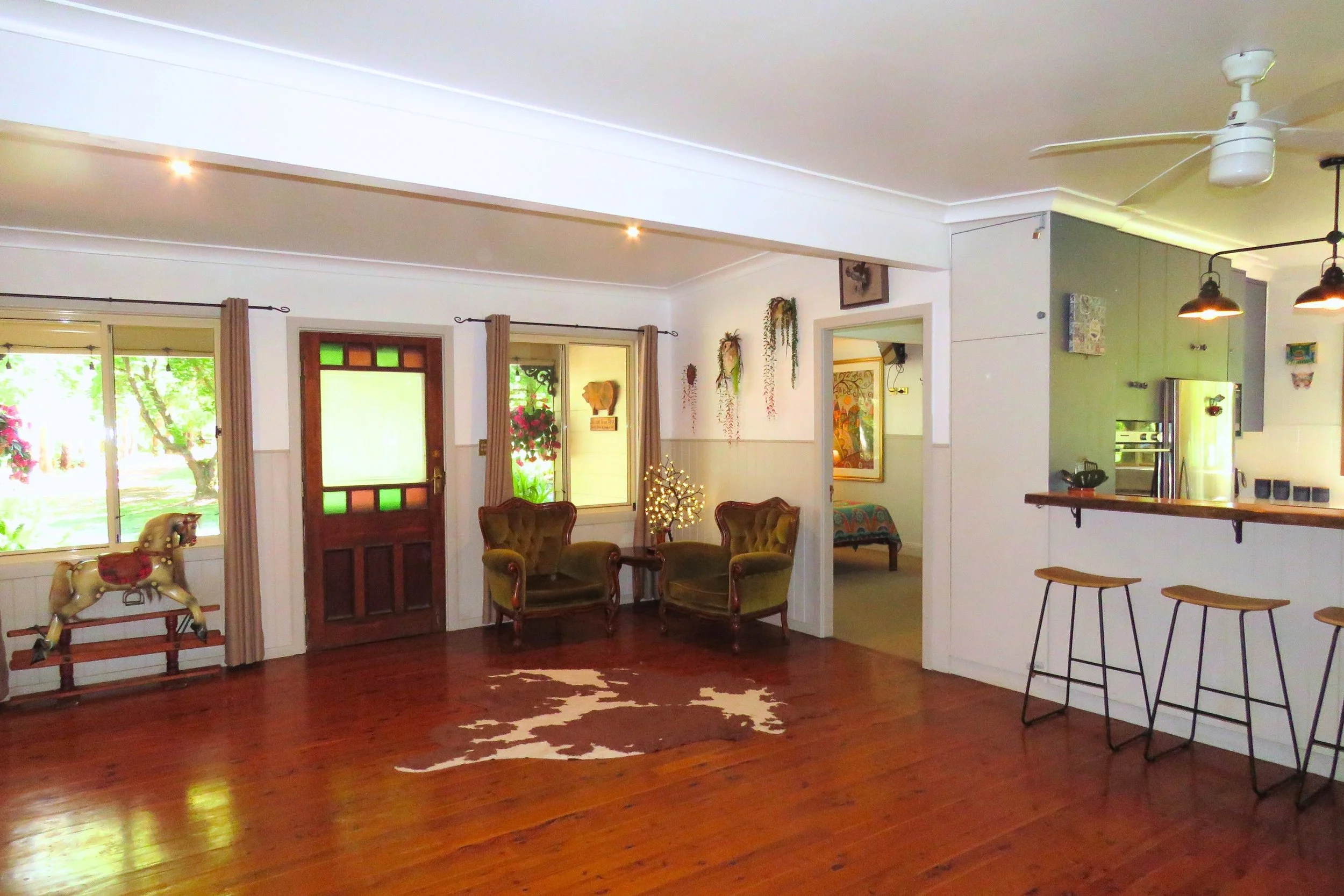 Living room with hardwood flooring, two green armchairs, a small side table with a plant, a cow-shaped rocking horse, large windows with curtains, and a front door with colored glass panels. An adjacent room with a colorful bed and artwork is visible