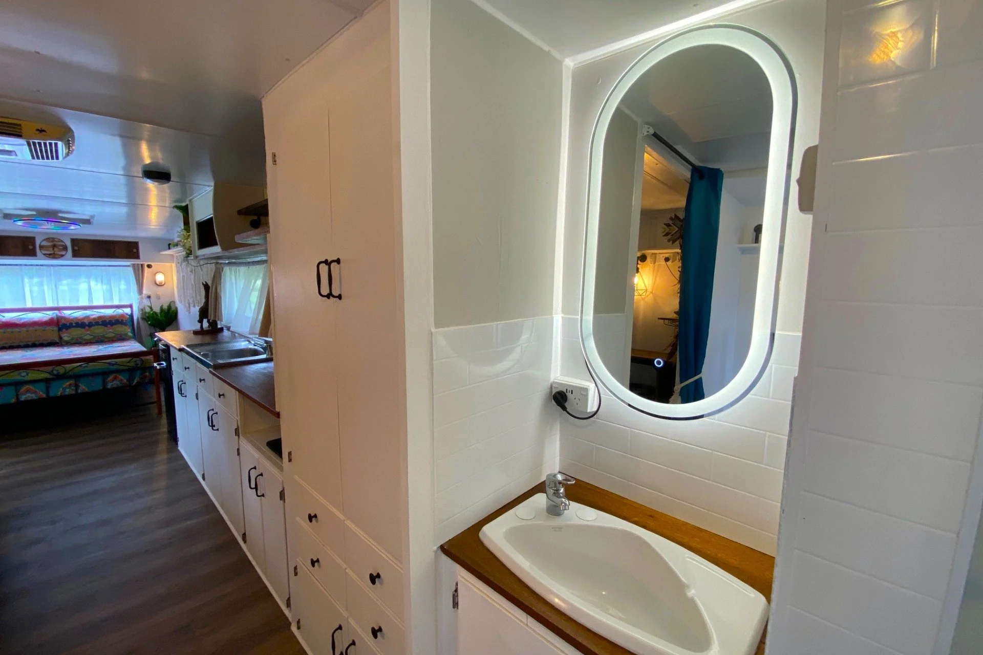 Interior of a small living space with a kitchenette and a sitting area, viewed through an open door or window. The space features a bed with colorful bedding, a window with white curtains, and wood flooring.