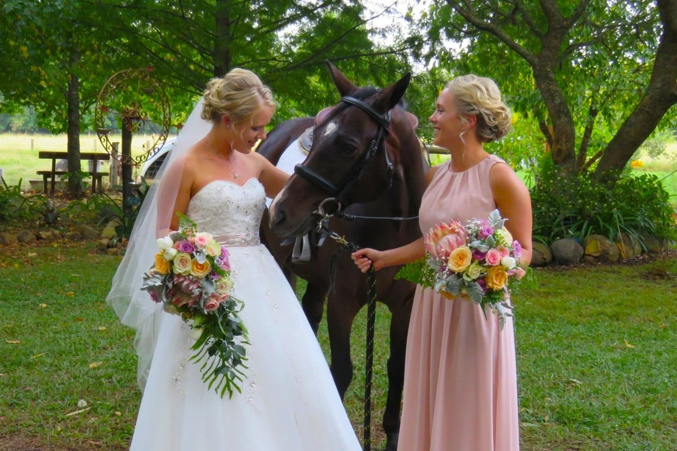 Two women in wedding attire holding bouquets stand next to a horse in a garden with trees and a bench in the background.