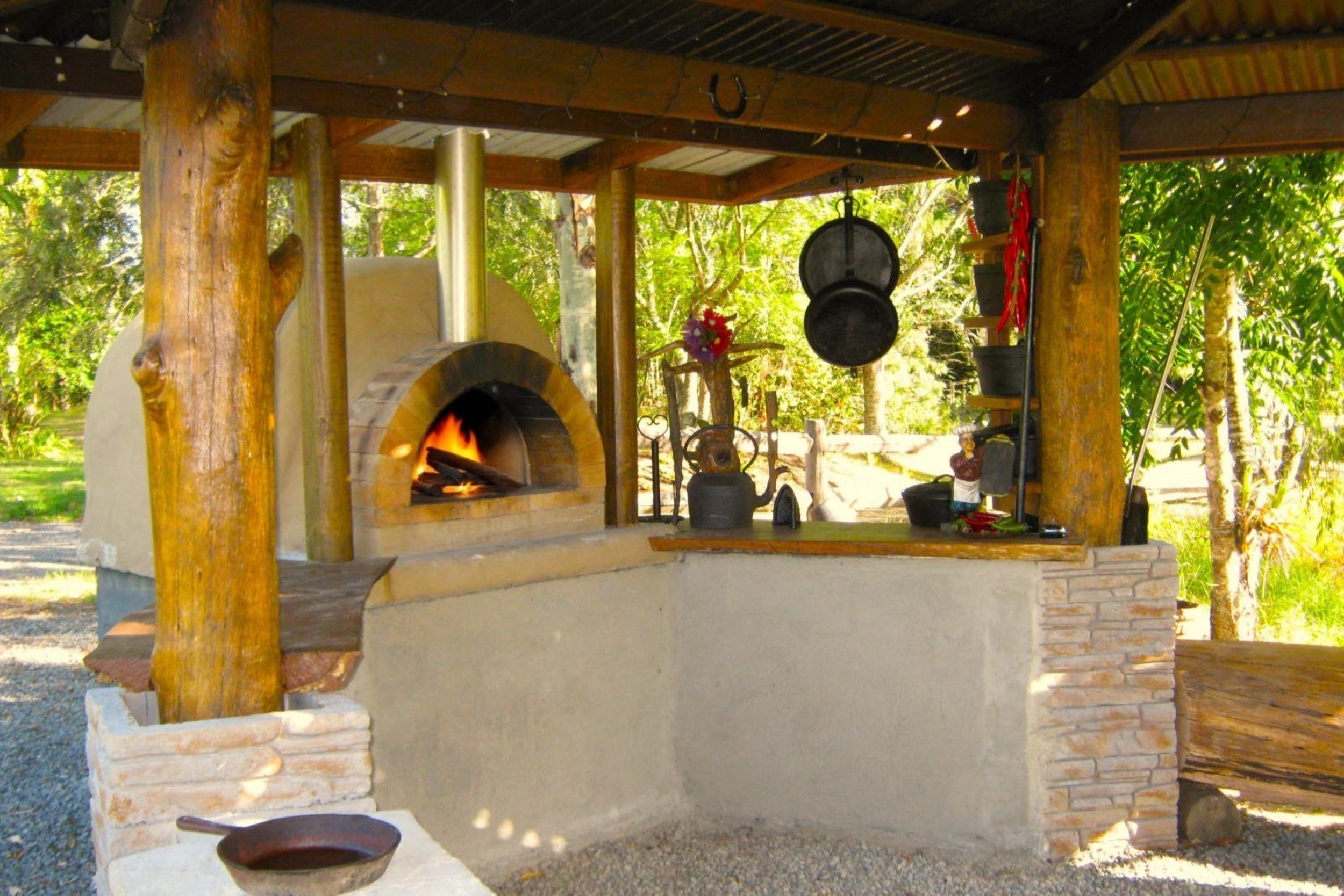 An outdoor traditional brick pizza oven with fire, under a wooden roof with hanging cast iron pots, in a lush green setting.
